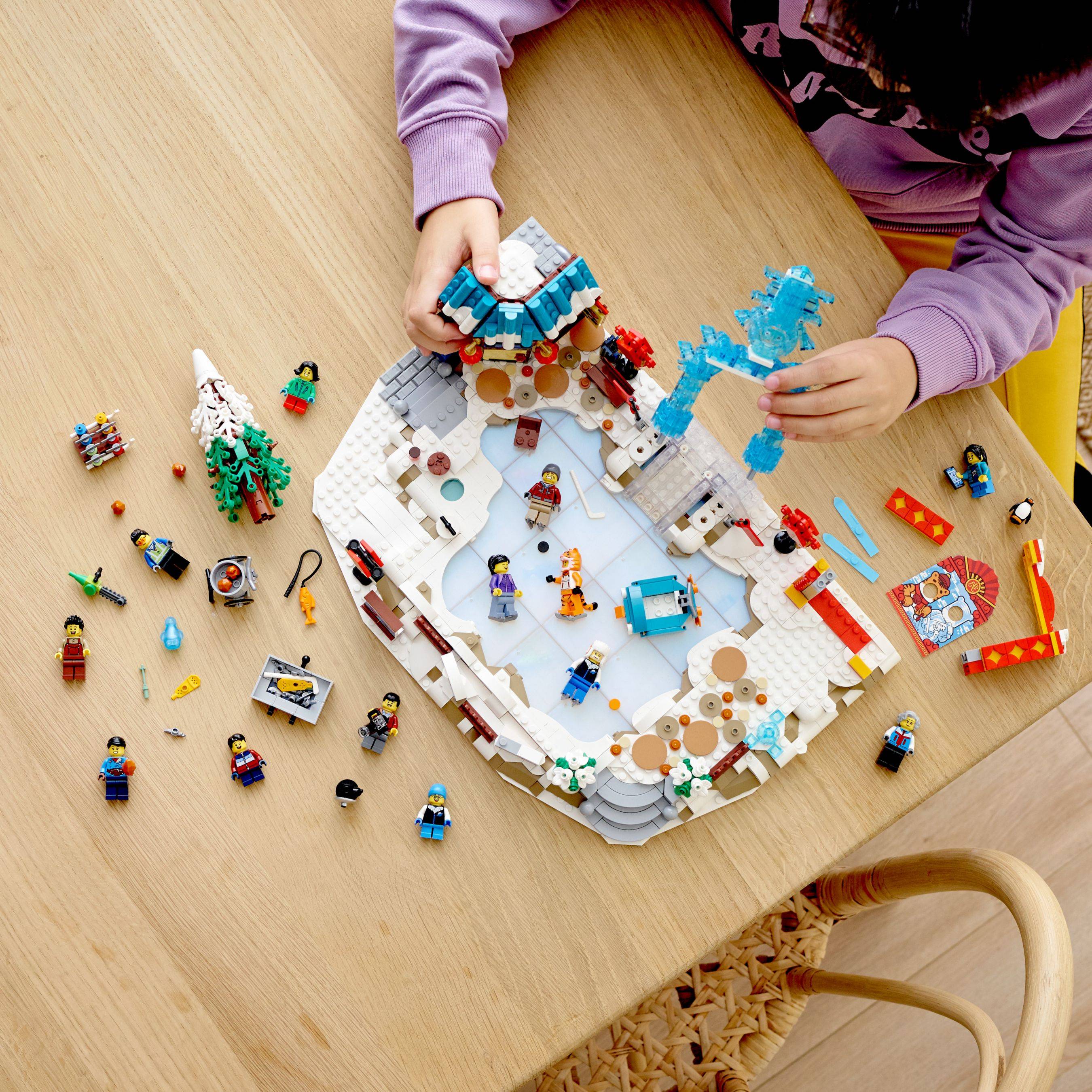 Kind baut eine LEGO-Winterdorfszene auf einem Tisch zusammen und platziert blaue Teile auf einem verschneiten kreisförmigen Bauwerk mit Minifiguren und verstreuten Teilen rundherum.