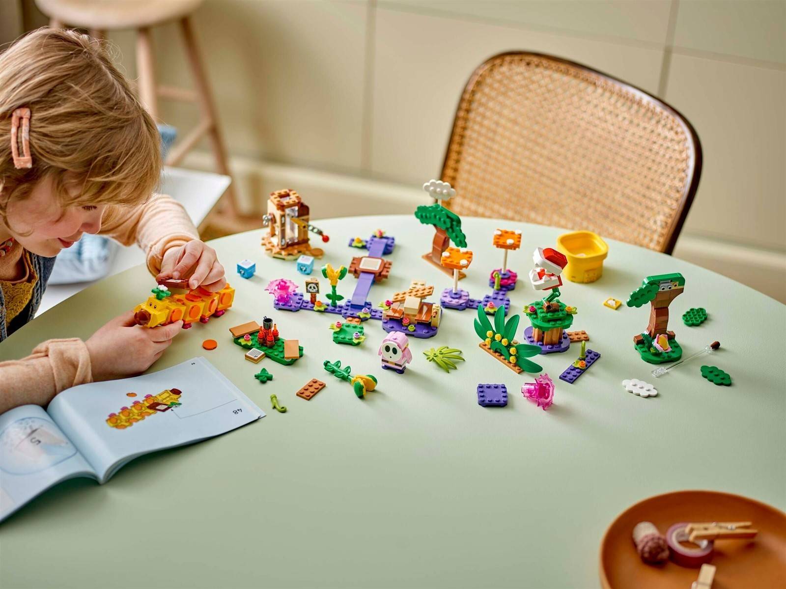 Ein Kind baut bunte LEGO-ähnliche Tier- und Pflanzenfiguren auf einem Tisch zusammen, während ein Anleitungsheft in der Nähe offen liegt.
