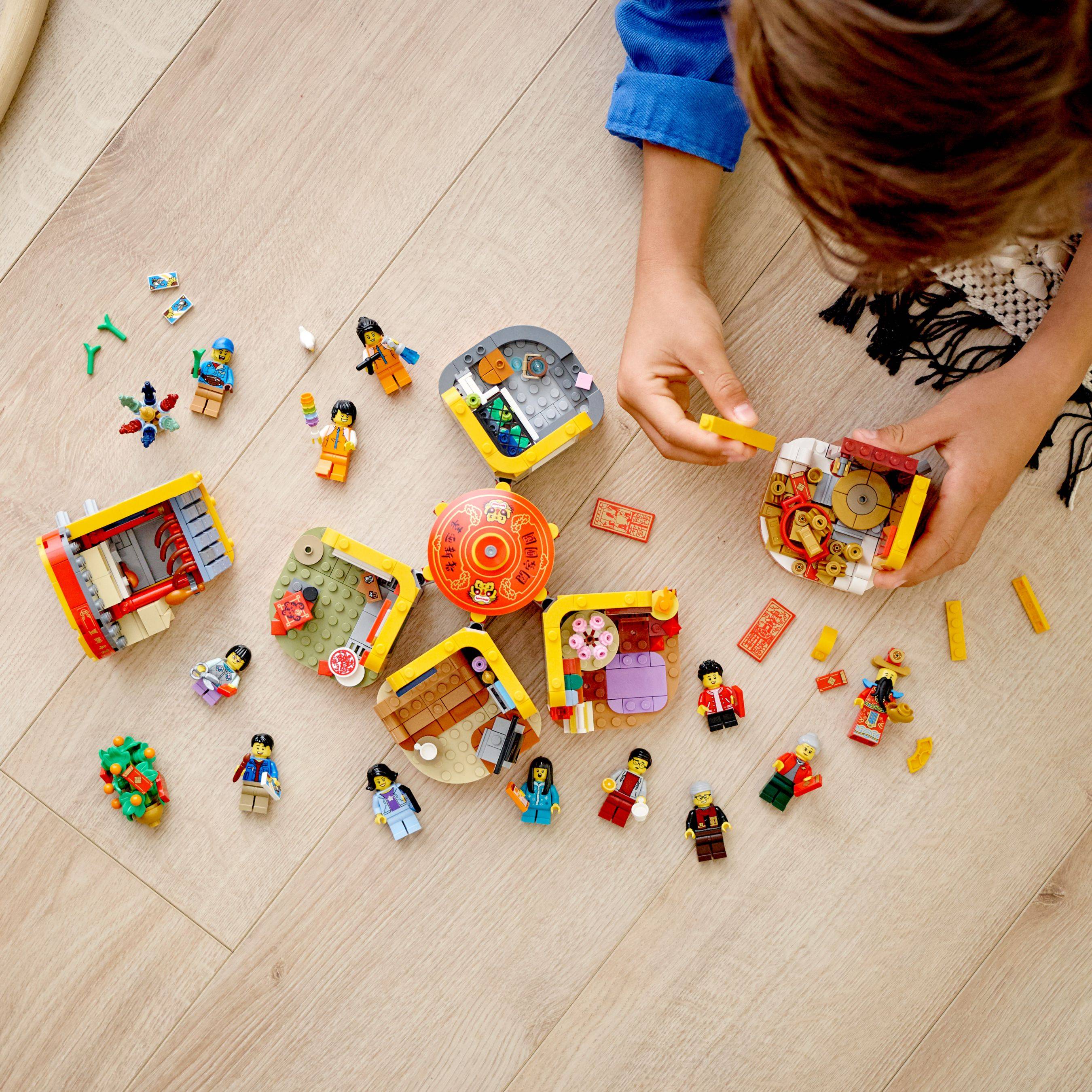 Draufsicht eines Kindes, das LEGO-ähnliche Miniaturgebäude und Figuren auf einem Holzboden zusammenbaut, mit vielen verstreuten losen Steinen und Teilen ringsum.