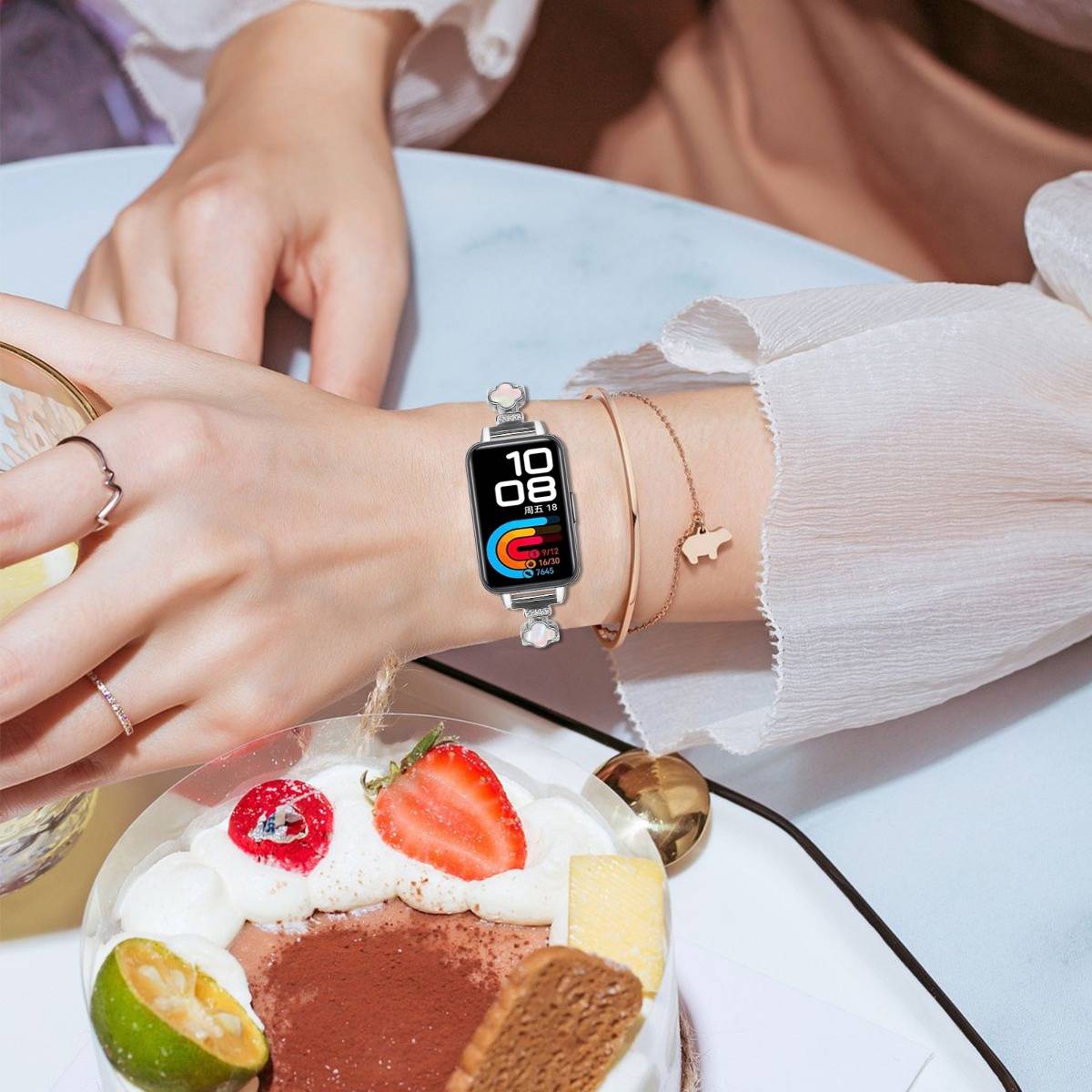 Nahaufnahme einer Smartwatch am Handgelenk einer Frau neben Dessert und Obst auf einem Tisch, zeigt die Uhrzeit 10:08 und farbige Aktivitätsringe.