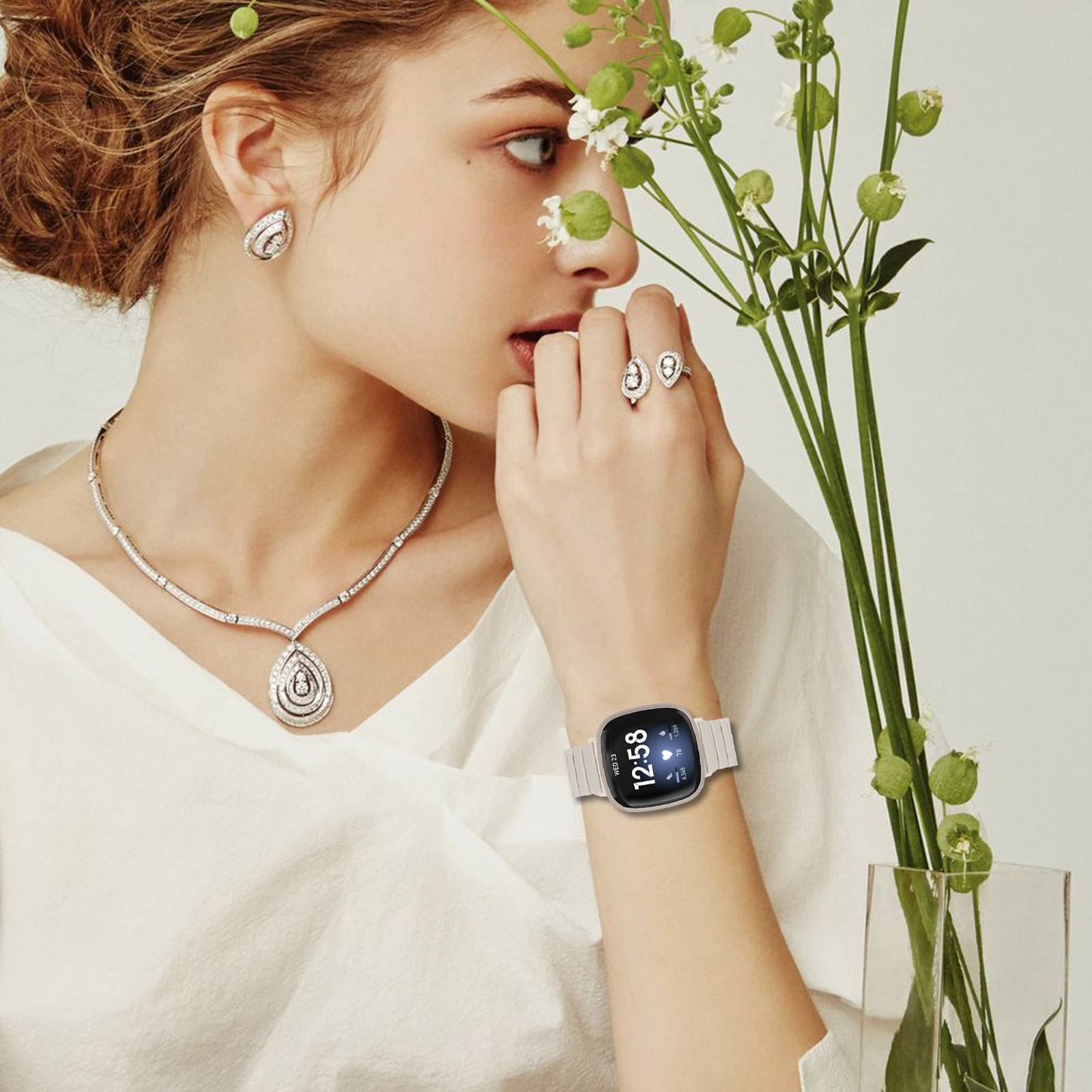Frau in weißem Top, silberner Schmuck tragend und eine Apple Watch tragend, neben grünen blühenden Stängeln posiert.