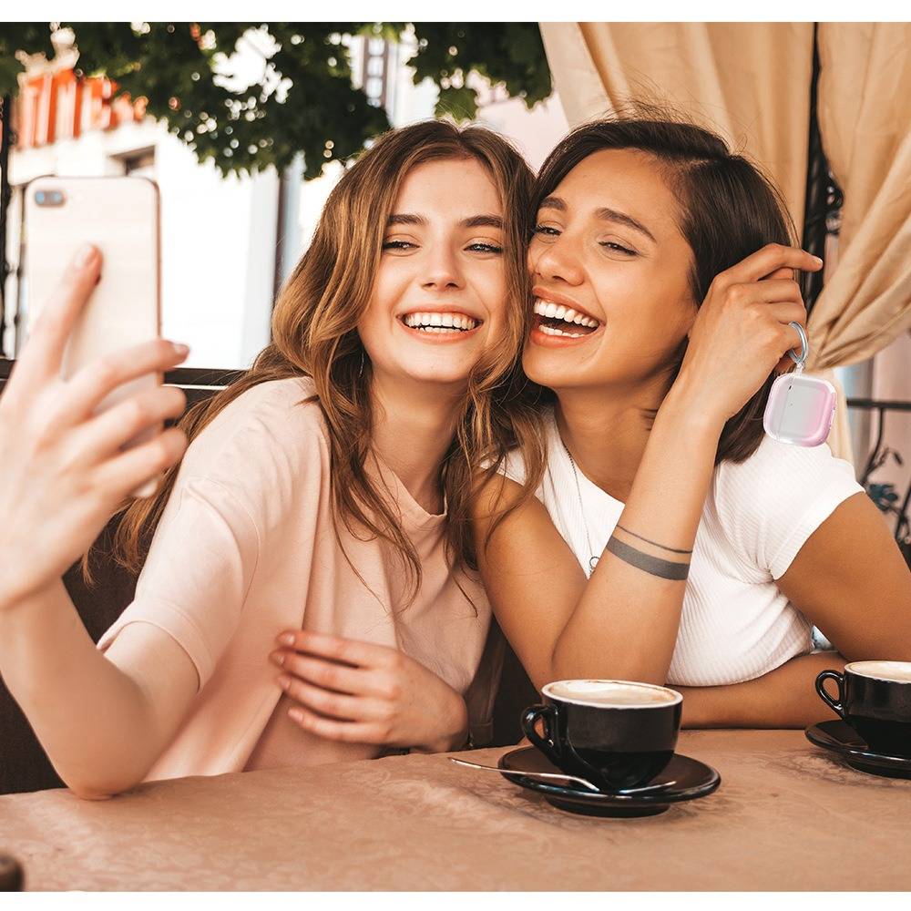 Zwei junge Frauen lächeln für ein Selfie an einem Cafétisch mit Kaffeetassen, eine hält ein Telefon im Vordergrund.
