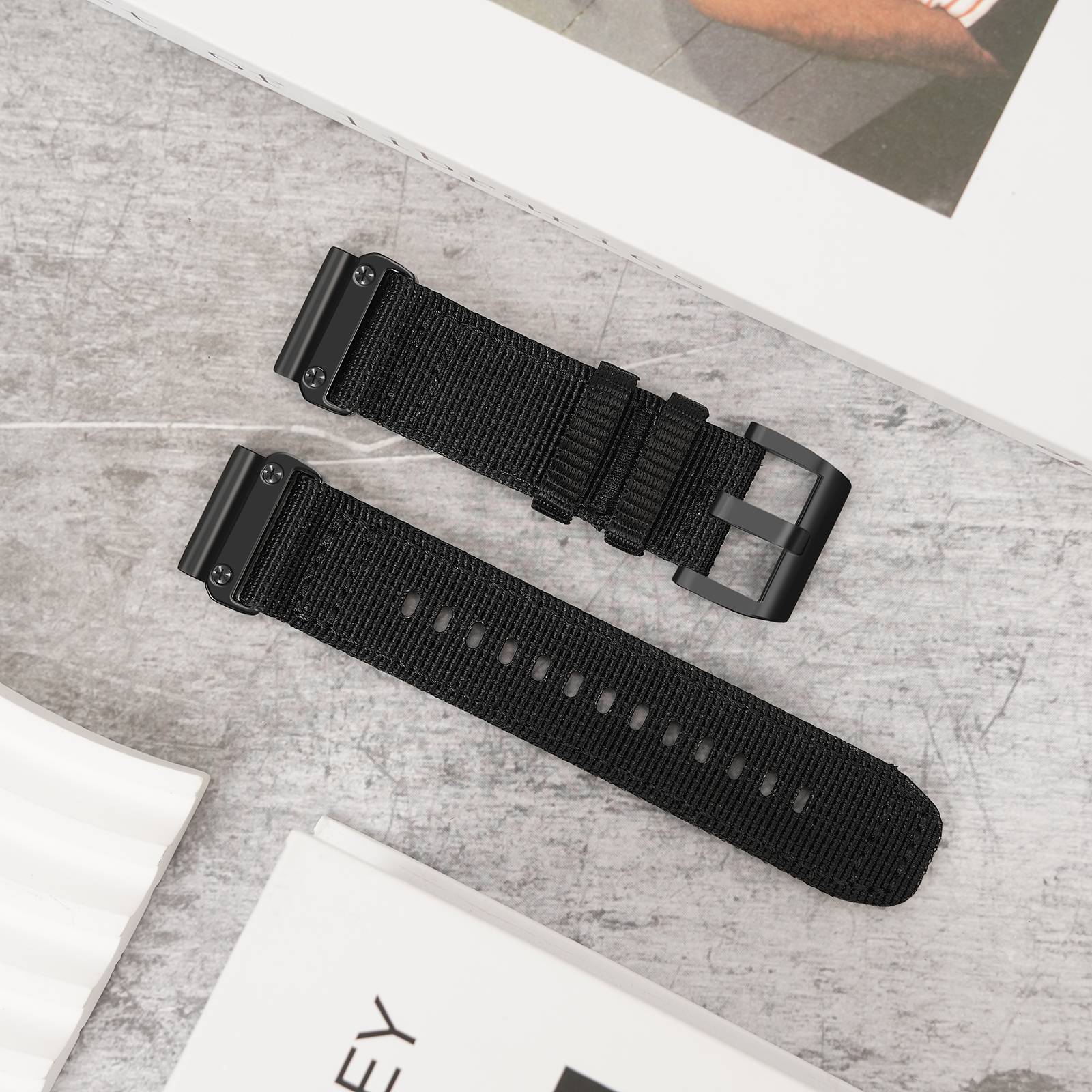 Zwei schwarze Nylon-Uhrenarmbänder mit mattem Metallschließen liegen diagonal auf einer grauen Oberfläche, dargestellt als Produktfotografie.
