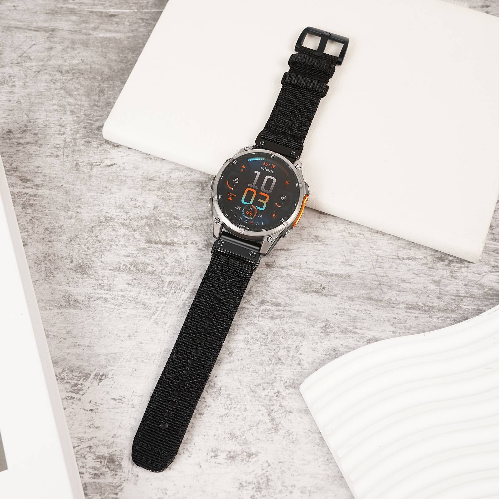 Schwarze runde digitale Armbanduhr mit langem Stoffarmband, diagonal auf einer hellgrauen Oberfläche neben weißen Blöcken gelegt.