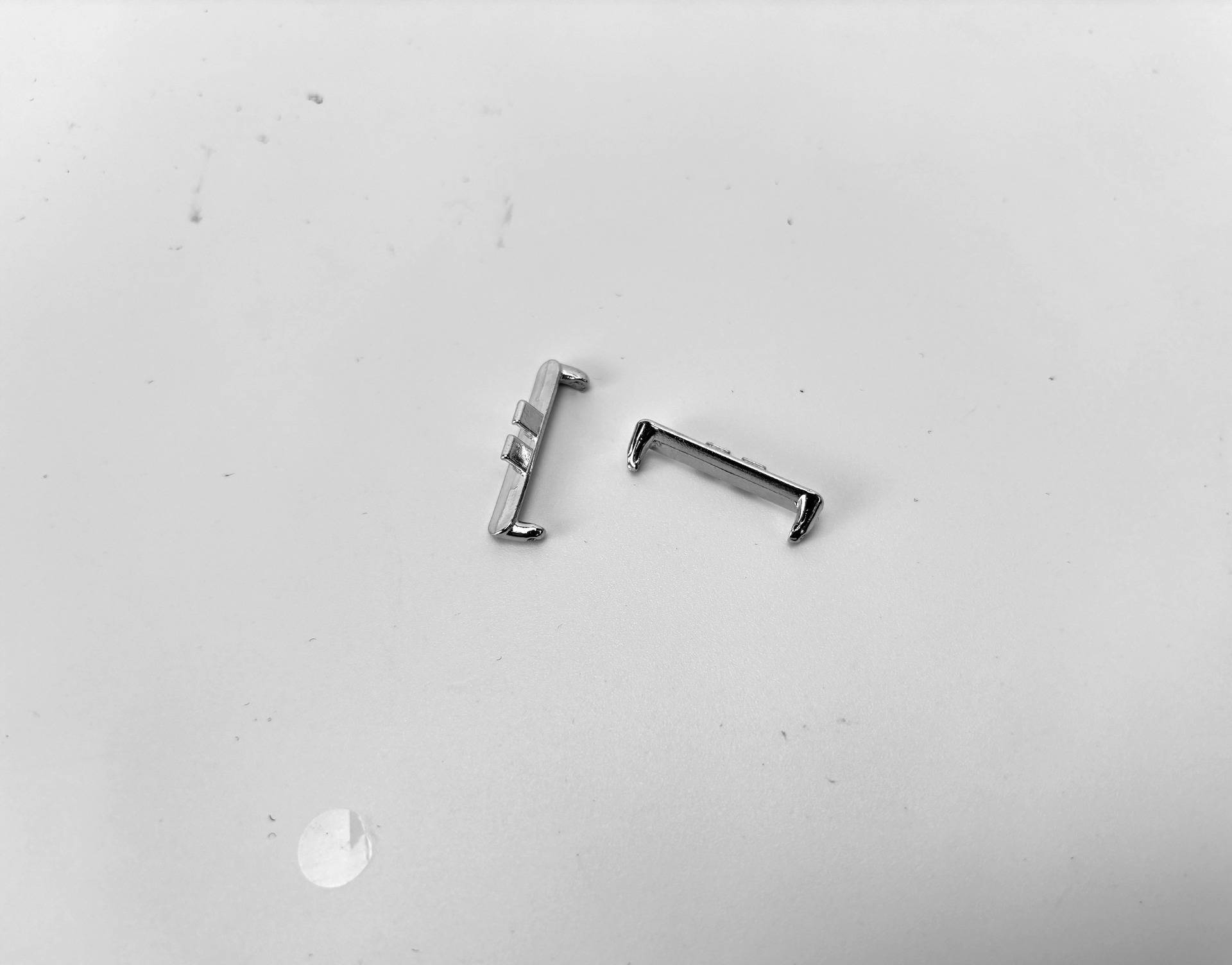 Zwei kleine Metallhardware-Clips oder Halterungen liegen auf einer einfachen, hellen Oberfläche, getrennt und in verschiedenen Winkeln angeordnet.