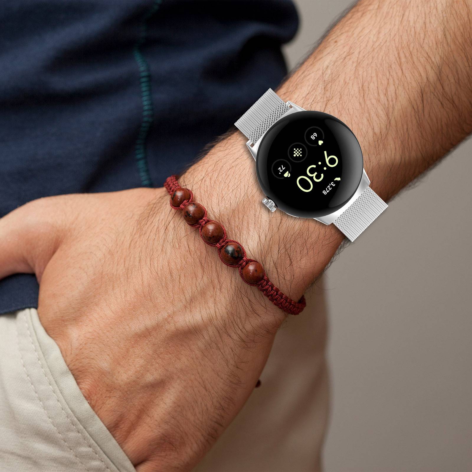 Nahaufnahme eines Unterarms mit einer runden Smartwatch mit silberfarbenem Mesh-Armband, das die Uhrzeit 9:30 anzeigt, neben einem roten Perlenarmband.