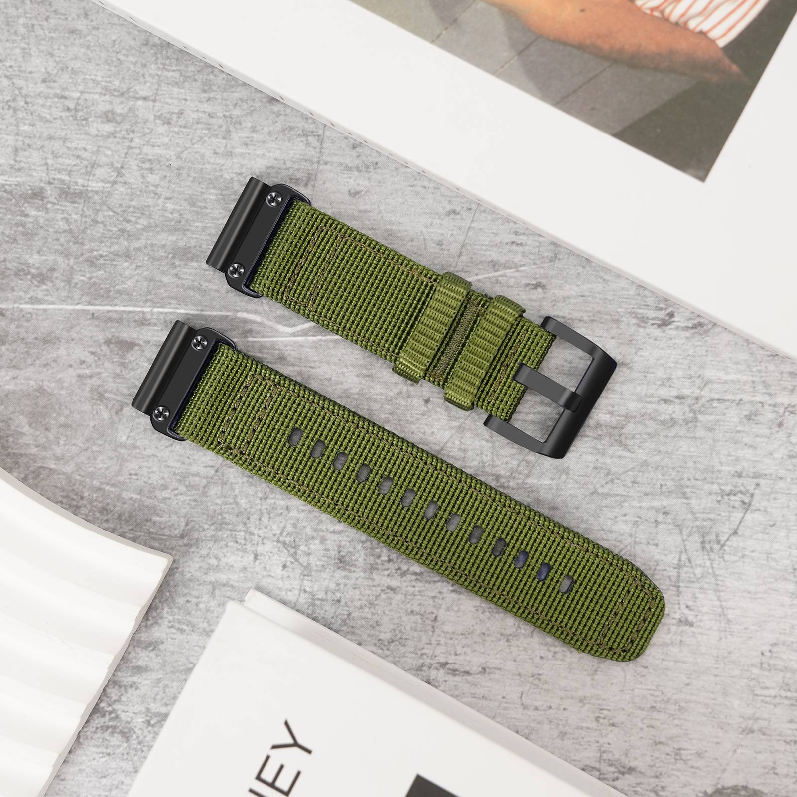 Zwei olivgrüne gewebte Uhrenarmbänder mit schwarzer Metallbeschlagnahme, eines mit Schnalle geschlossen und eines offen, auf einer grauen Oberfläche ausgestellt.