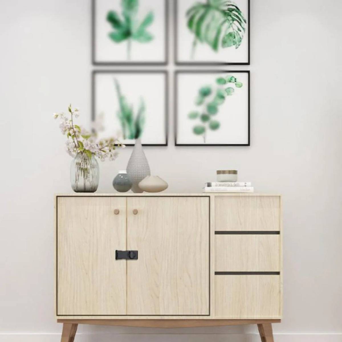 Minimalistischer Schrank aus hellem Holz mit Vasen und Büchern, gekrönt von einem Blumenarrangement, unter vier gerahmten grünen botanischen Drucken an einer weißen Wand.