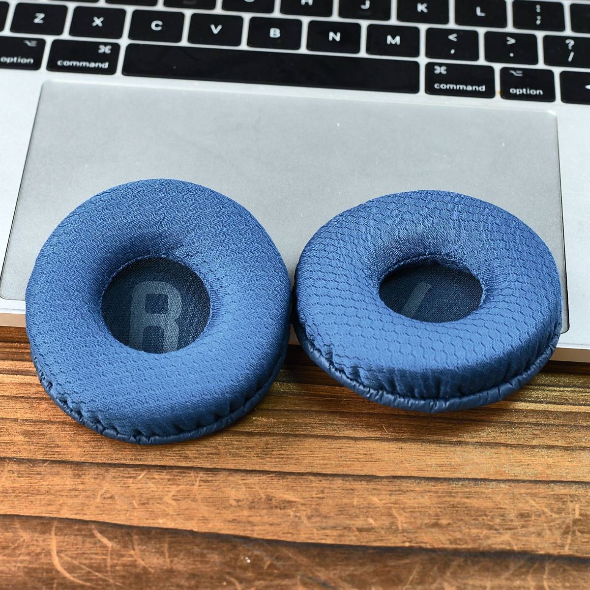 Zwei blaue Ersatz-Ohrpolster liegen auf einem Holzschreibtisch vor einer Laptop-Tastatur.