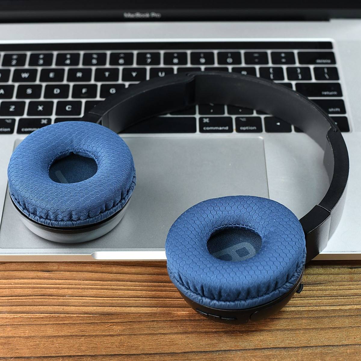 Blaue gepolsterte Over-Ear-Kopfhörer, die auf einer silbernen Laptop-Tastatur und einem Trackpad ruhen, auf einer Holztischplatte.