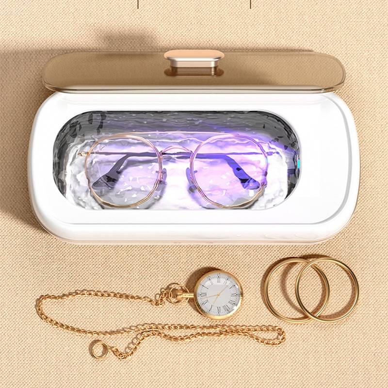 Brille in einem UV-Desinfektionskasten mit goldenem Schmuck darunter, einschließlich einer Uhr und Ketten, auf einem beigefarbenen Stoffhintergrund.