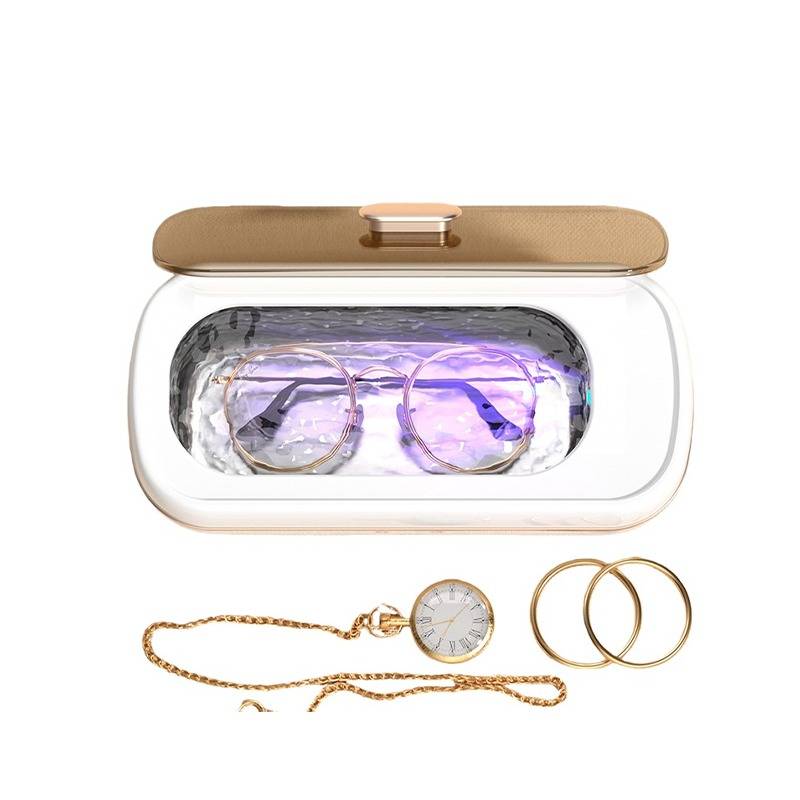 Durchsichtige Brillenetui mit goldenem Deckel und violettem Innenfutter, dargestellt über einer goldenen Kettenarmbanduhr und zwei goldenen Creolen auf weißem Hintergrund.
