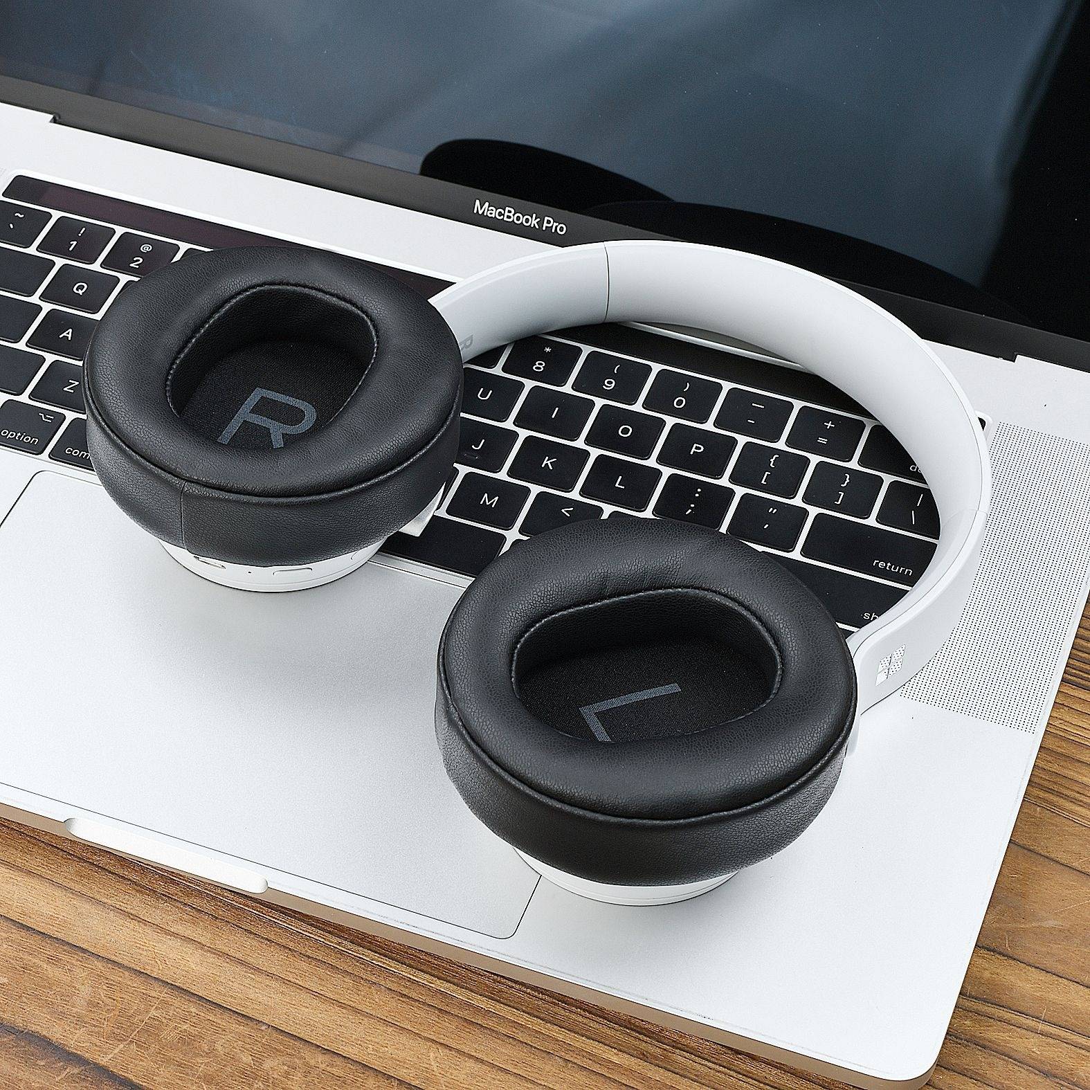 Weiße Over-Ear-Kopfhörer mit schwarzen Ohrpolstern, die auf einer MacBook-Pro-Tastatur und einem Trackpad auf einem Holzschreibtisch ruhen.