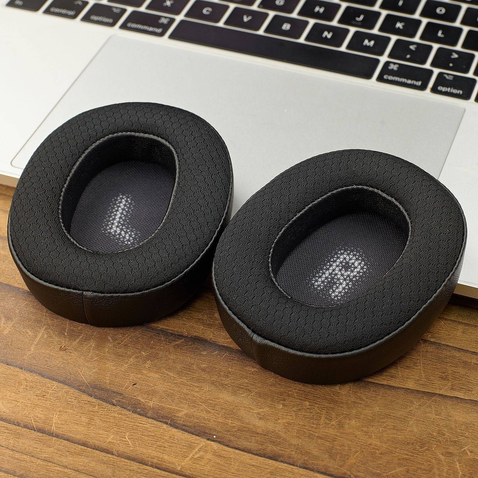 Zwei schwarze ovale Ohrpolster für Over-Ear-Kopfhörer ruhen auf einem Holzschreibtisch vor einer Laptop-Tastatur.