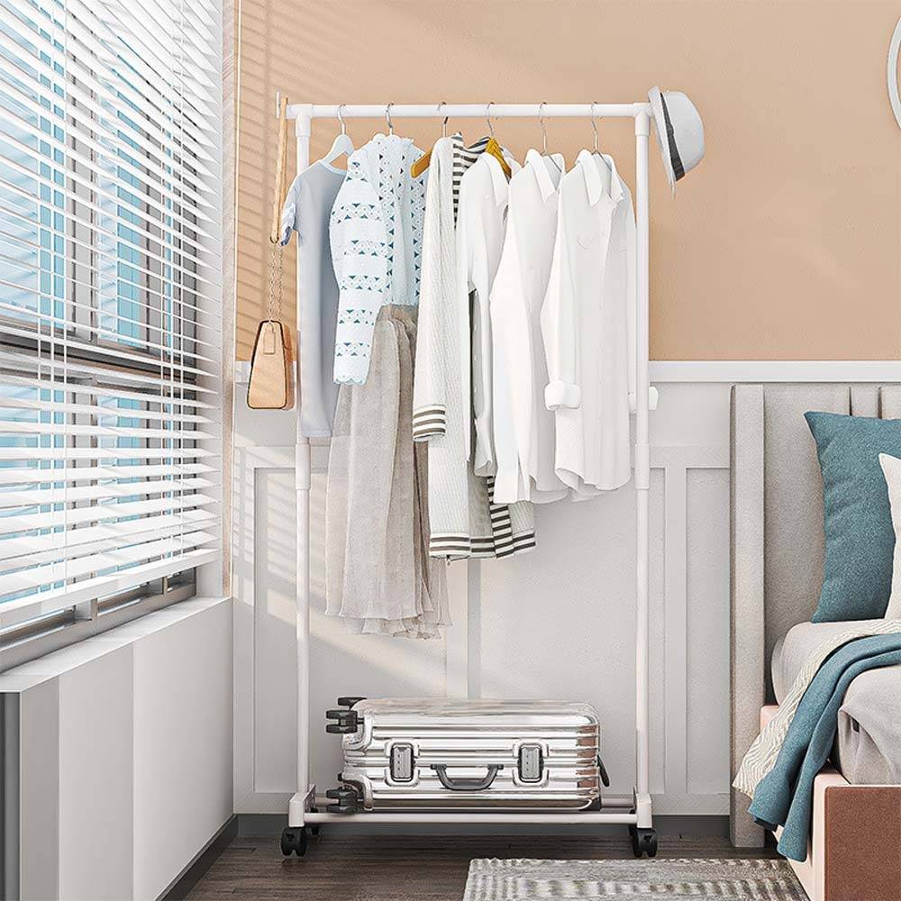 Weiße Kleiderstange mit hängenden Kleidungsstücken und einem Hut neben einem Fenster, neben einem Bett in einem hellen Schlafzimmer.