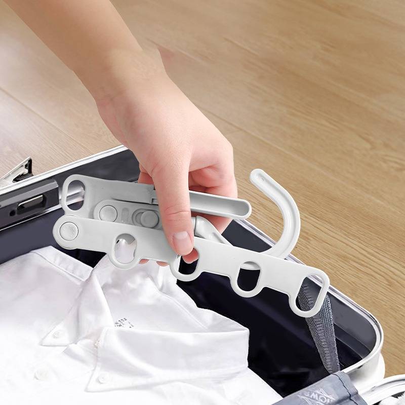 Hand, die einen zusammenklappbaren Kleiderbügel mit mehreren Haken in einen offenen Koffer mit gefalteter weißer Kleidung legt.