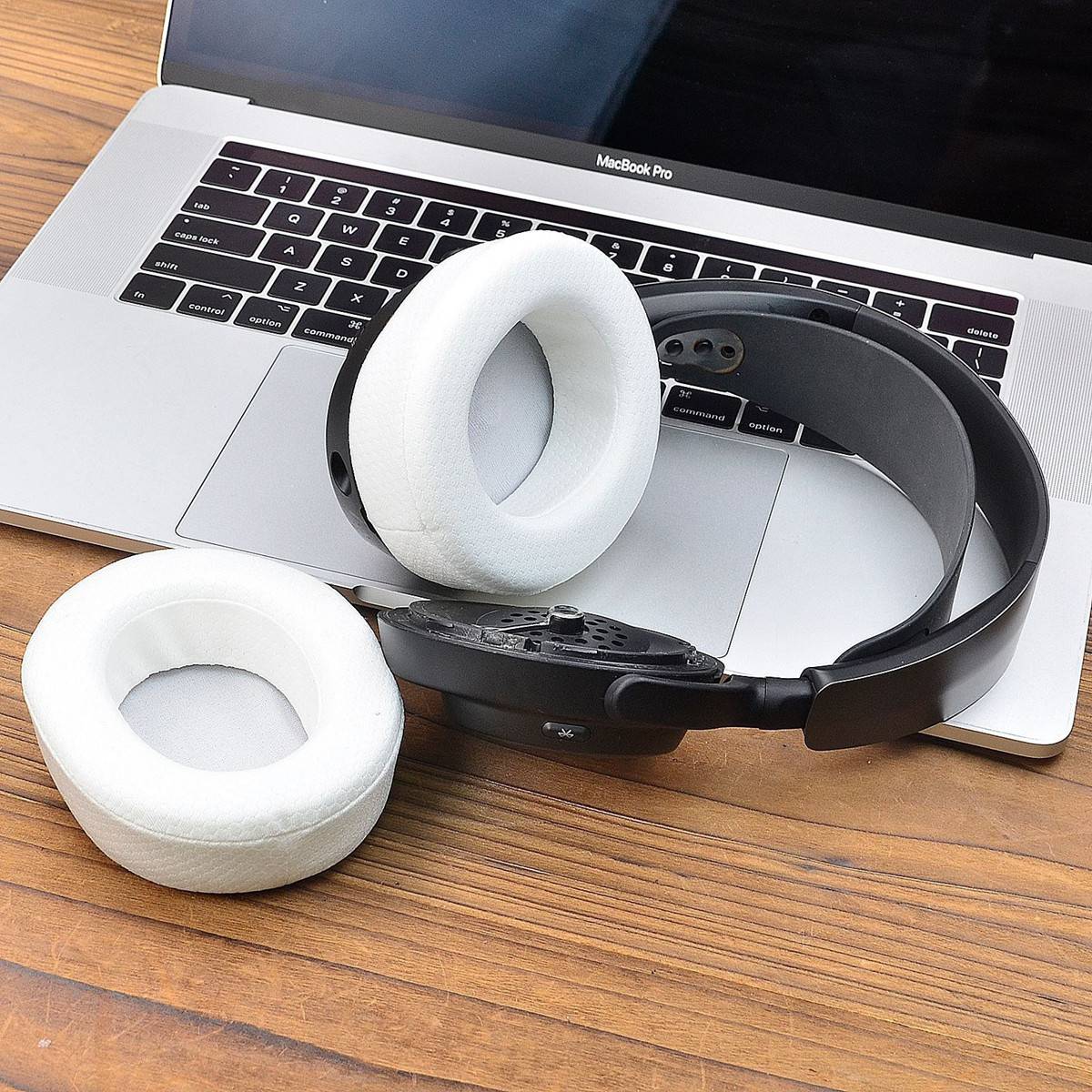 Drei runde weiße Ohrpolster und ein schwarzes Kopfbügel liegen auf einer MacBook-Pro-Tastatur auf einem Holzschreibtisch und deuten auf zerlegt Kopfhörer hin.