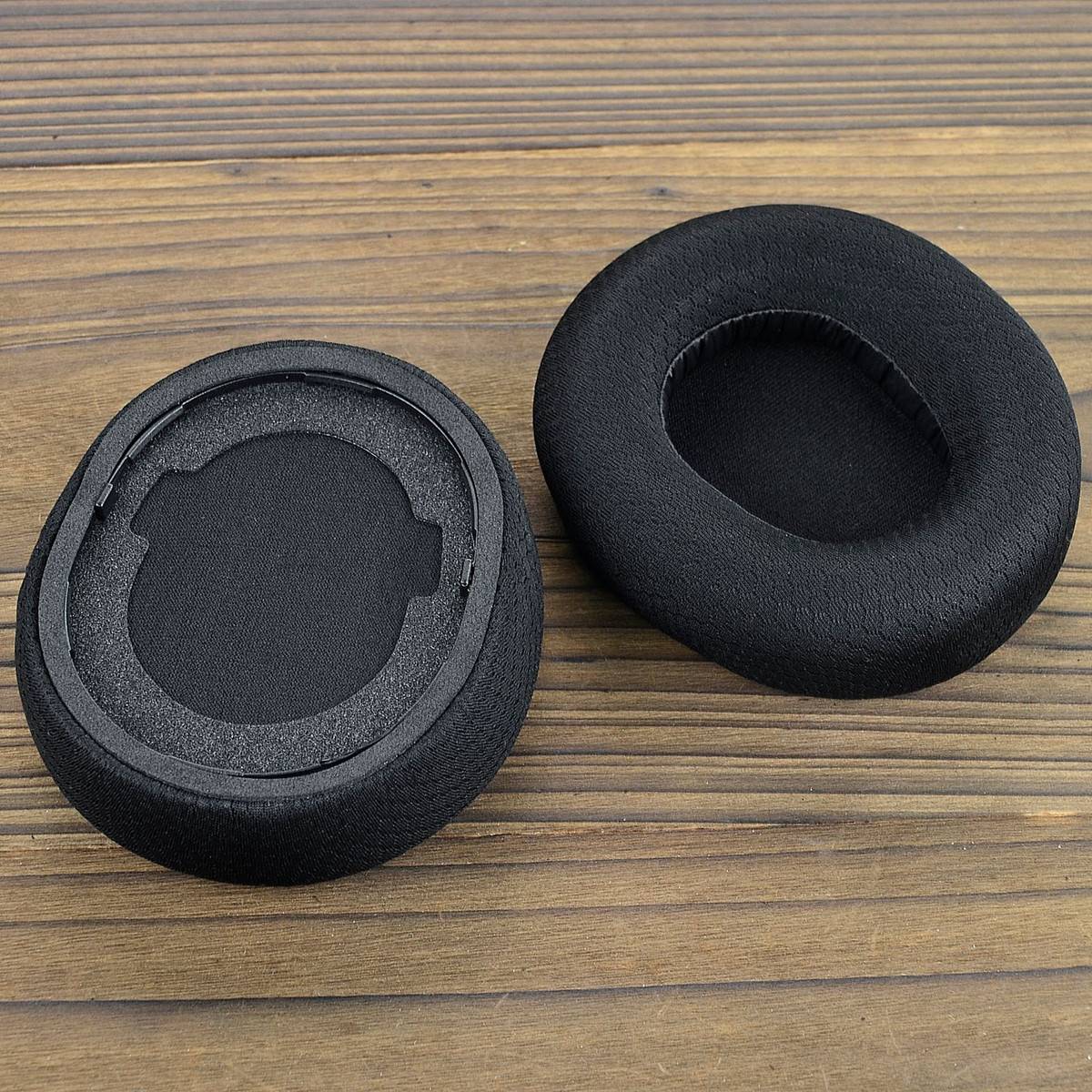 Zwei schwarze runde Ohrmuscheln auf einer Holzoberfläche, eine zeigt den Befestigungsring auf der Unterseite und die andere die gepolsterte Außenseite.