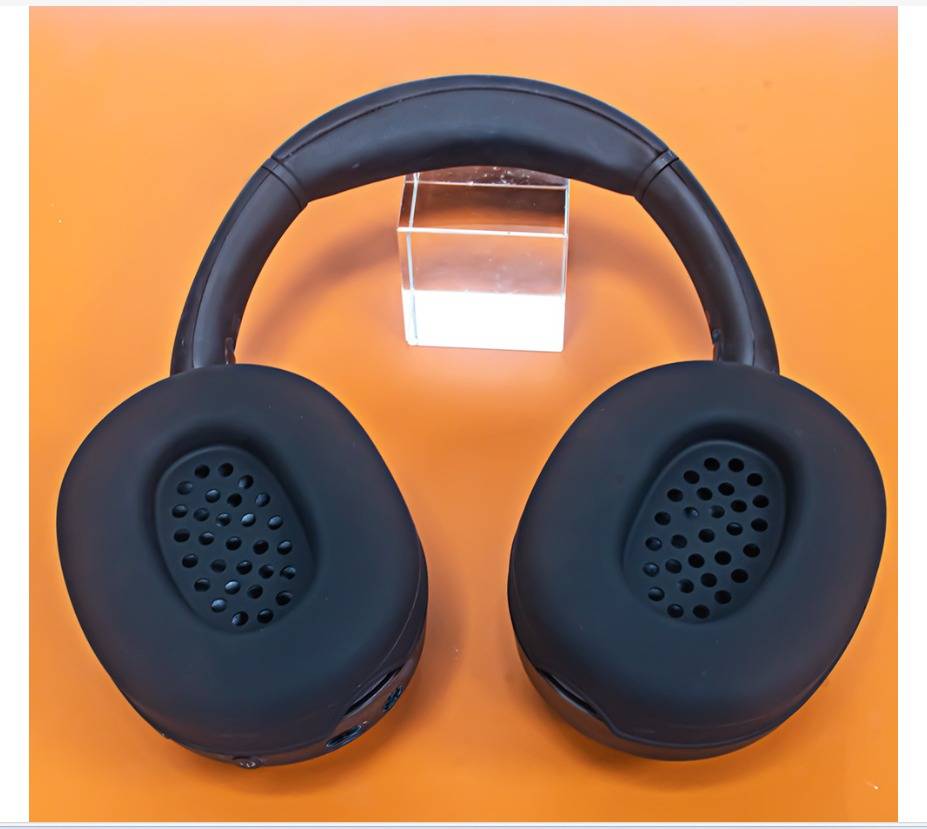 Schwarze Over-Ear-Kopfhörer auf orangefarbenem Hintergrund dargestellt, Frontalansicht mit durchsichtigem Ständer hinter dem Kopfbügel.