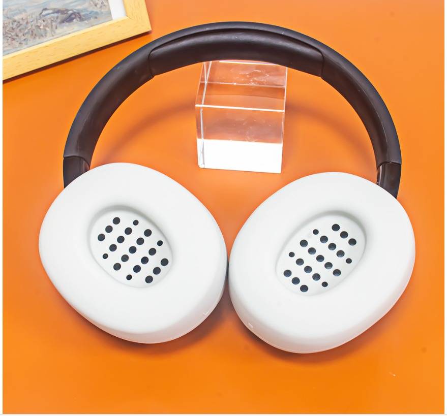 Weiße Over-Ear-Kopfhörer mit schwarzem Kopfbügel auf einer orangefarbenen Oberfläche, gezeigt mit einem durchsichtigen Displayständer dahinter.