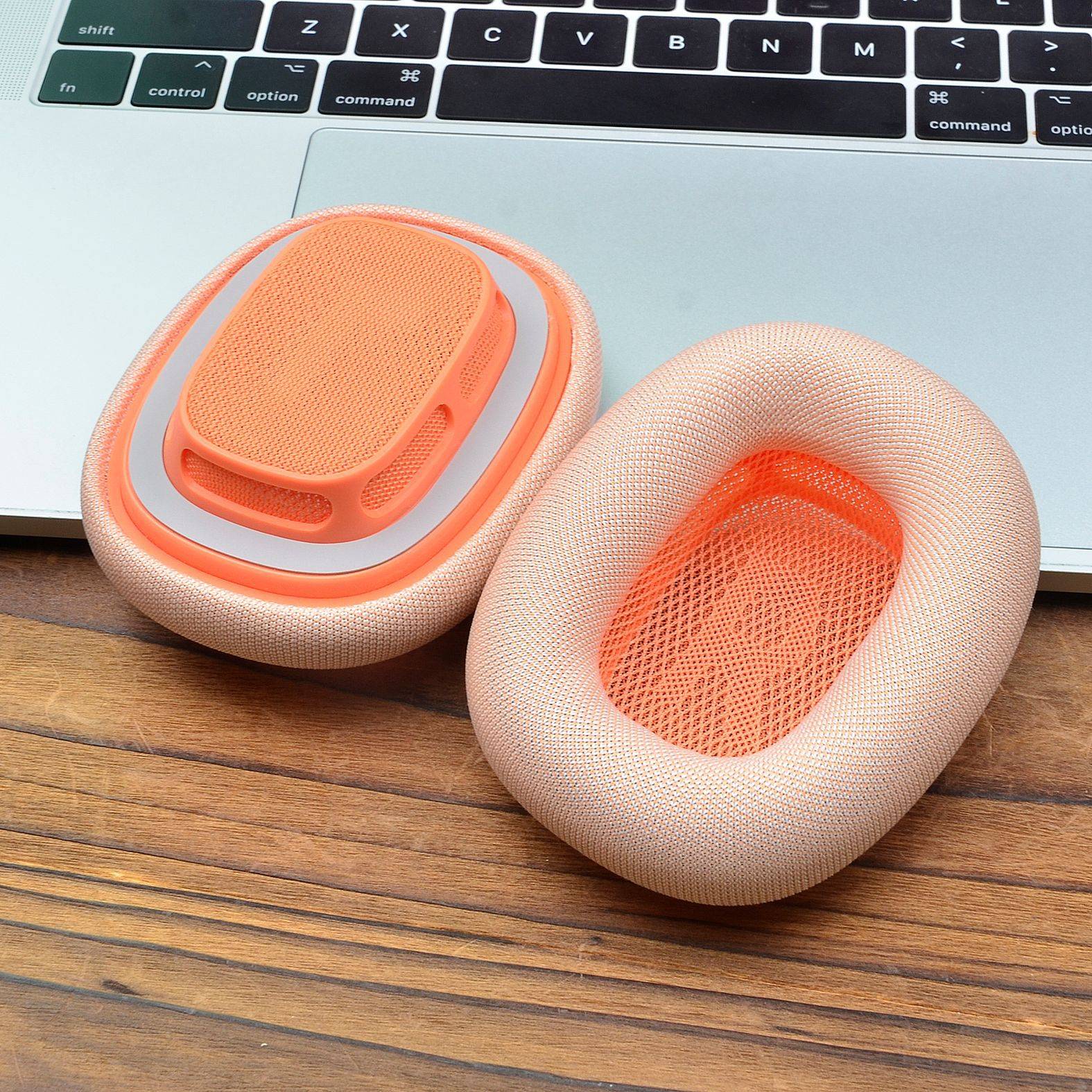 Zwei orange-und-beige Mesh-Ohrpolster oder tragbare Pads liegen auf einem Holzschreibtisch neben einer Laptop-Tastatur.