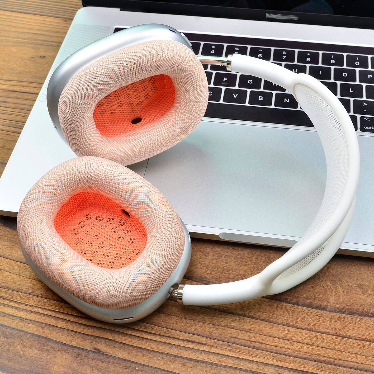 Weiße Over-Ear-Kopfhörer mit korallenfarbenen Ohrpolstern liegen auf einer Laptop-Tastatur auf einem Holzschreibtisch.
