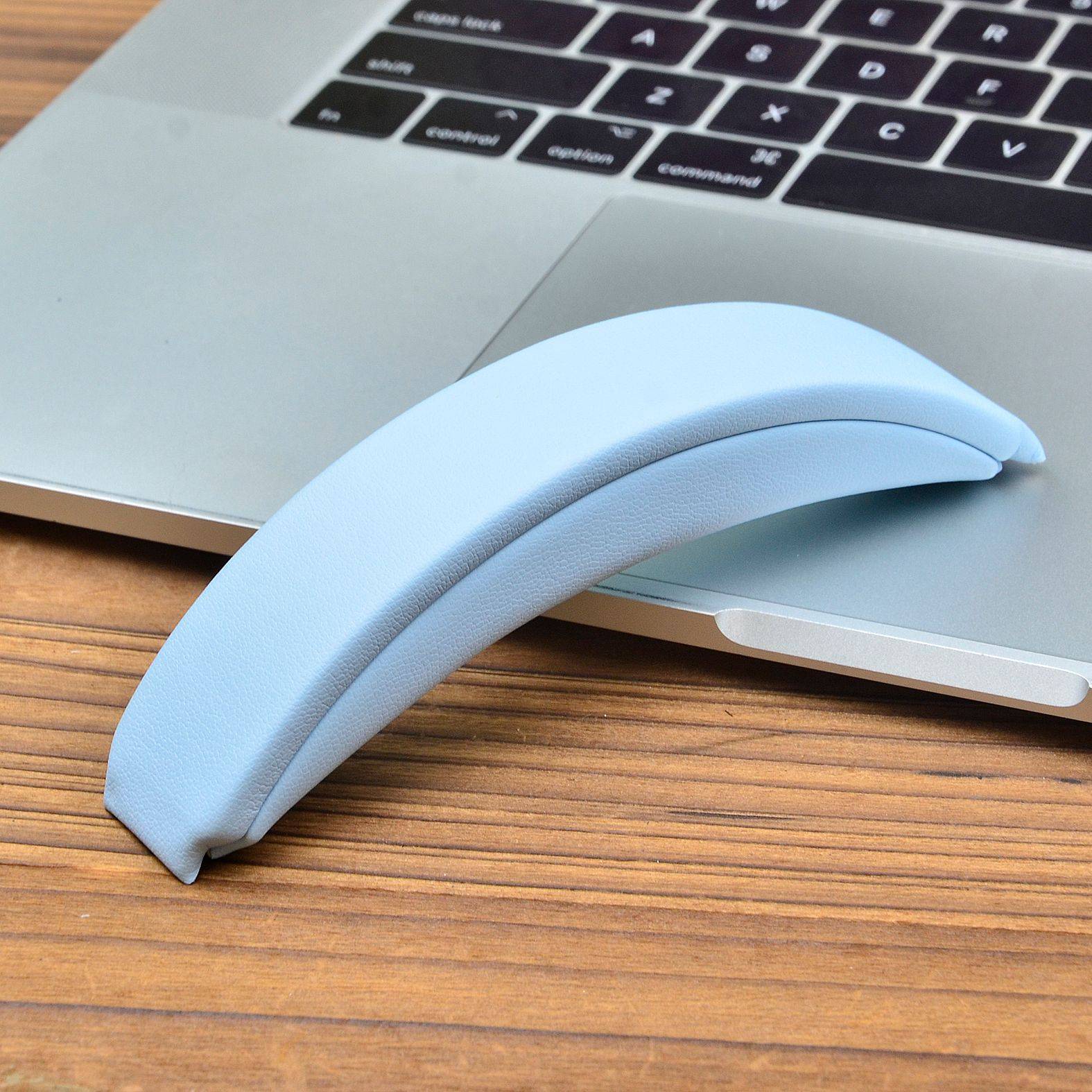 Eine hellblaue ergonomische Handgelenkauflage, die an der Kante eines Laptops auf einem Holzschreibtisch angelehnt ist.