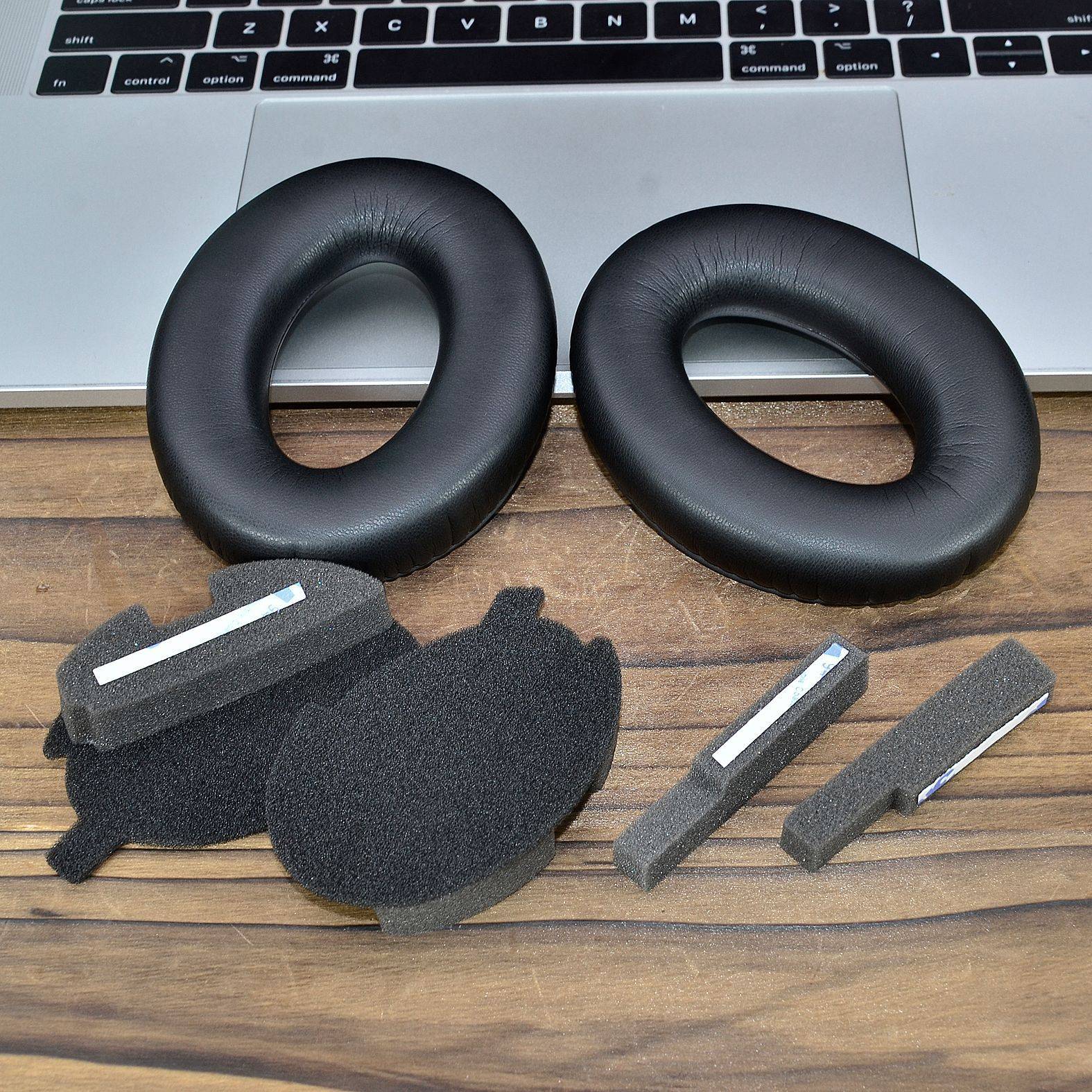 Zwei schwarze Ersatzohrpolster und Schaumstoffüberzüge für Over-Ear-Kopfhörer werden auf einem Holztisch vor einem Laptop gezeigt.