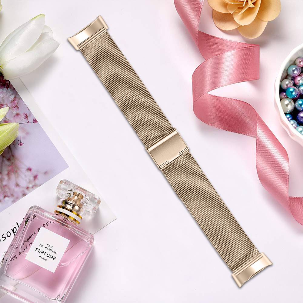 Goldfarbenes Mesh-Uhrenarmband auf einer pastellfarbenen Flachlage mit rosafarbener Parfümflasche, Band, Blumen und Perlen.