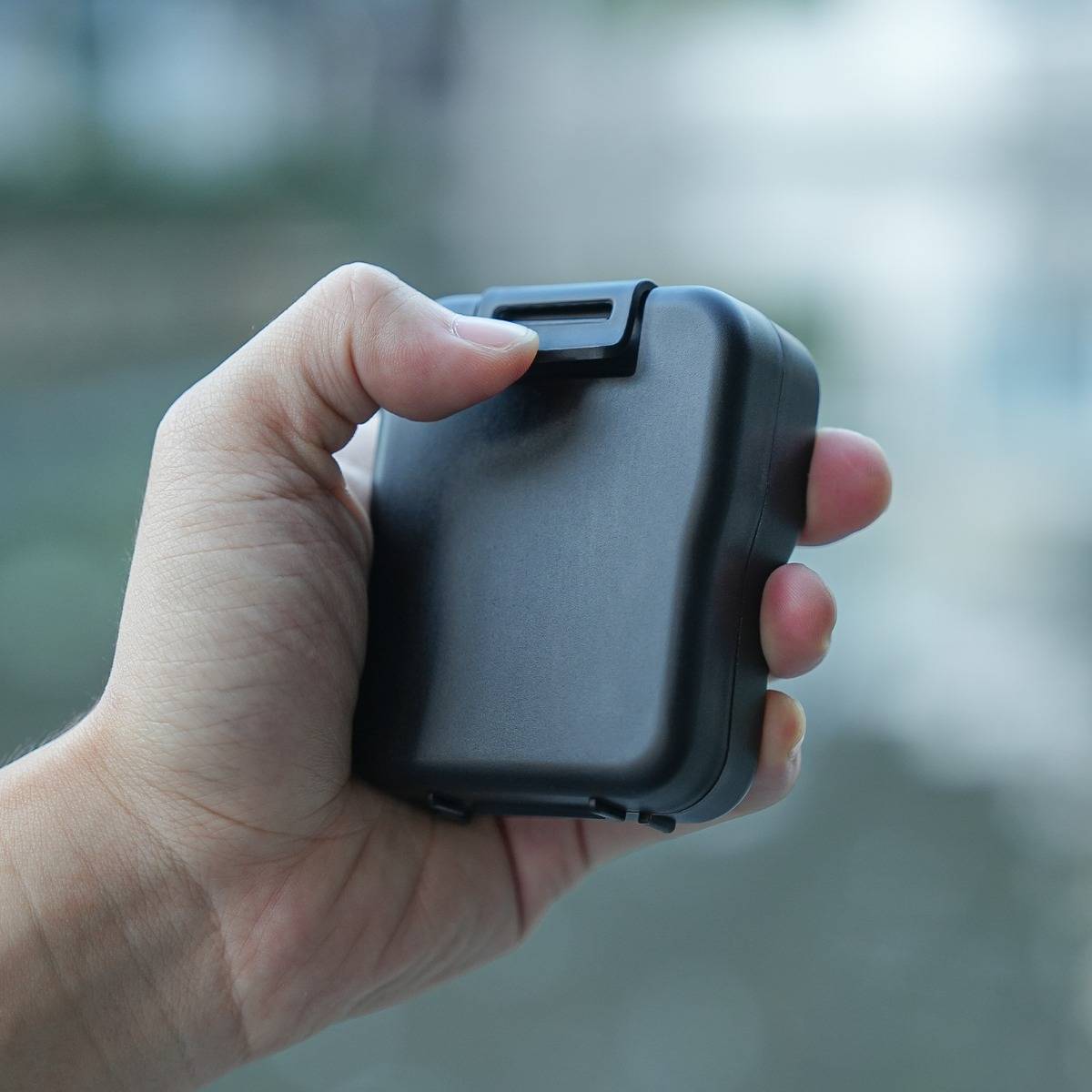 Hand, die ein kleines schwarzes rechteckiges Gerät mit einem Clip oder einer Verriegelung oben hält, vor einem unscharfen Außenhintergrund.