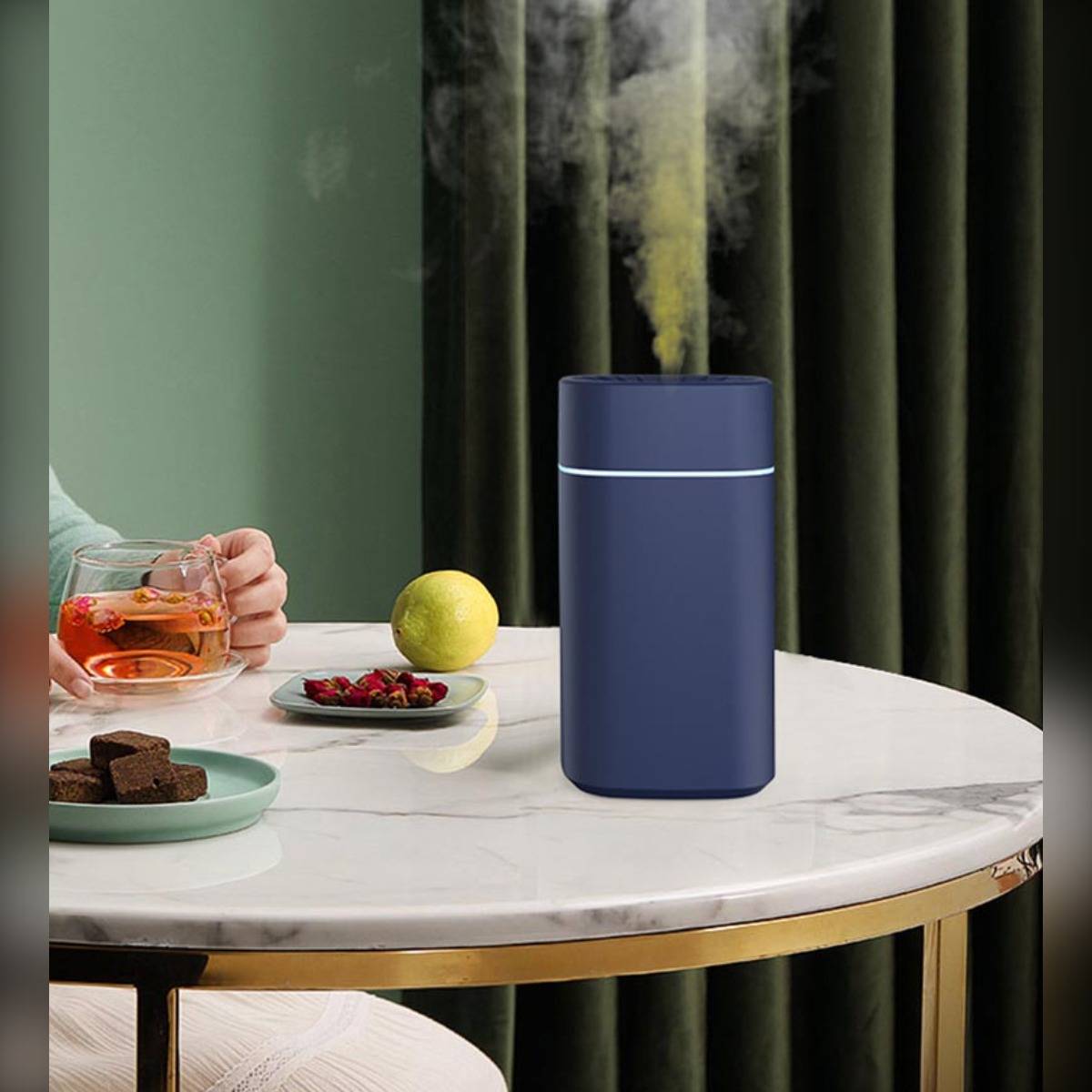 Blauer zylindrischer Luftbefeuchter auf einem Marmortisch, der Dampf neben Tee, einer Zitrone und Snacks in einer gemütlichen Inneneinrichtung abgibt.