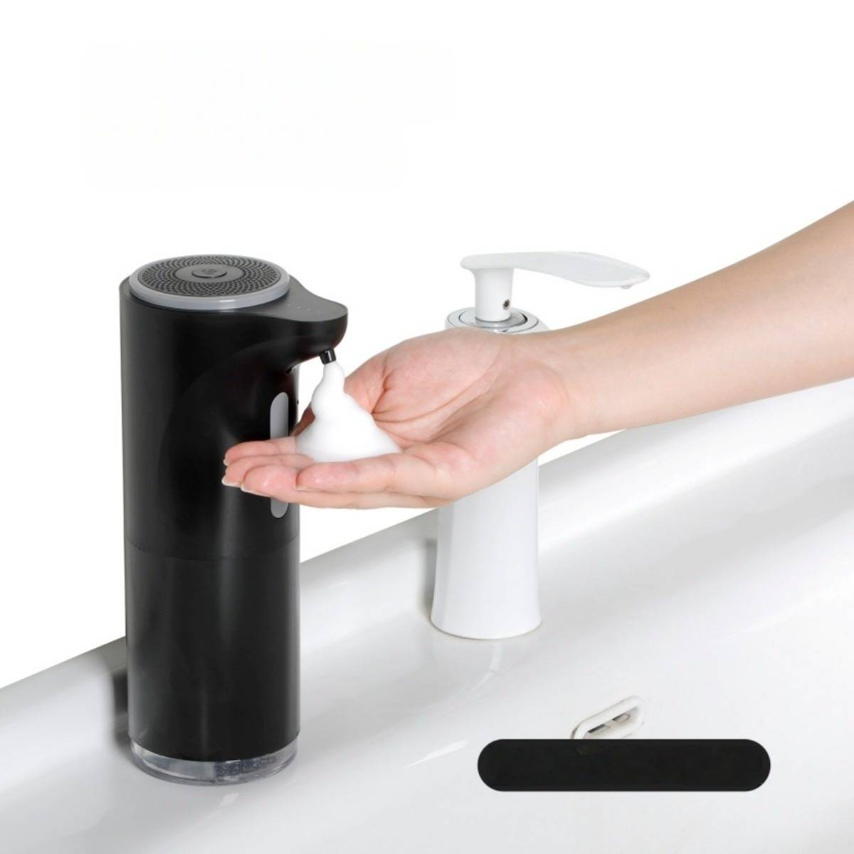 Hand dispensiert Schaumseife aus einem schwarzen Pumpspender neben einem weißen Spender auf einer Spülentheke.