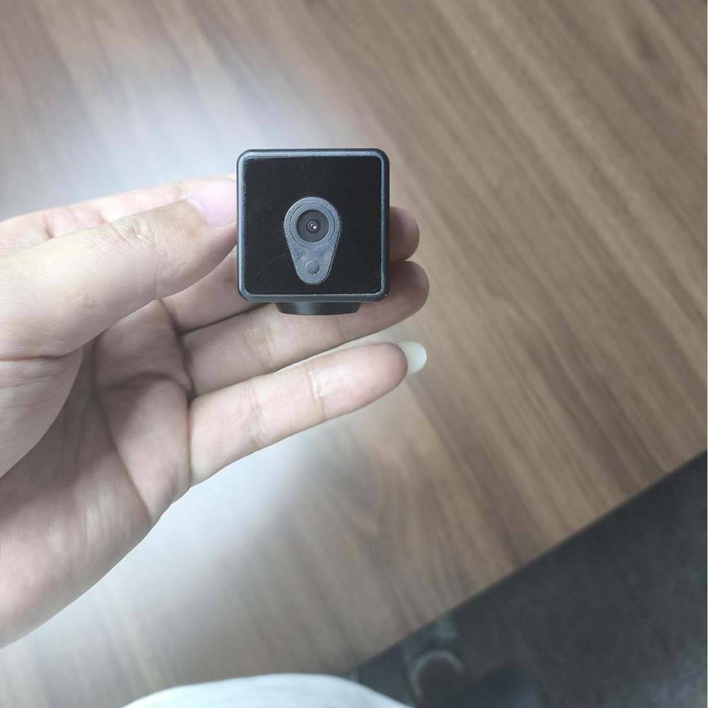 Eine Hand, die eine kleine schwarze quadratische Kamera oder einen Sensor mit einem einzelnen Objektiv hält, in Nahaufnahme vor einem Holzboden-Hintergrund gezeigt.