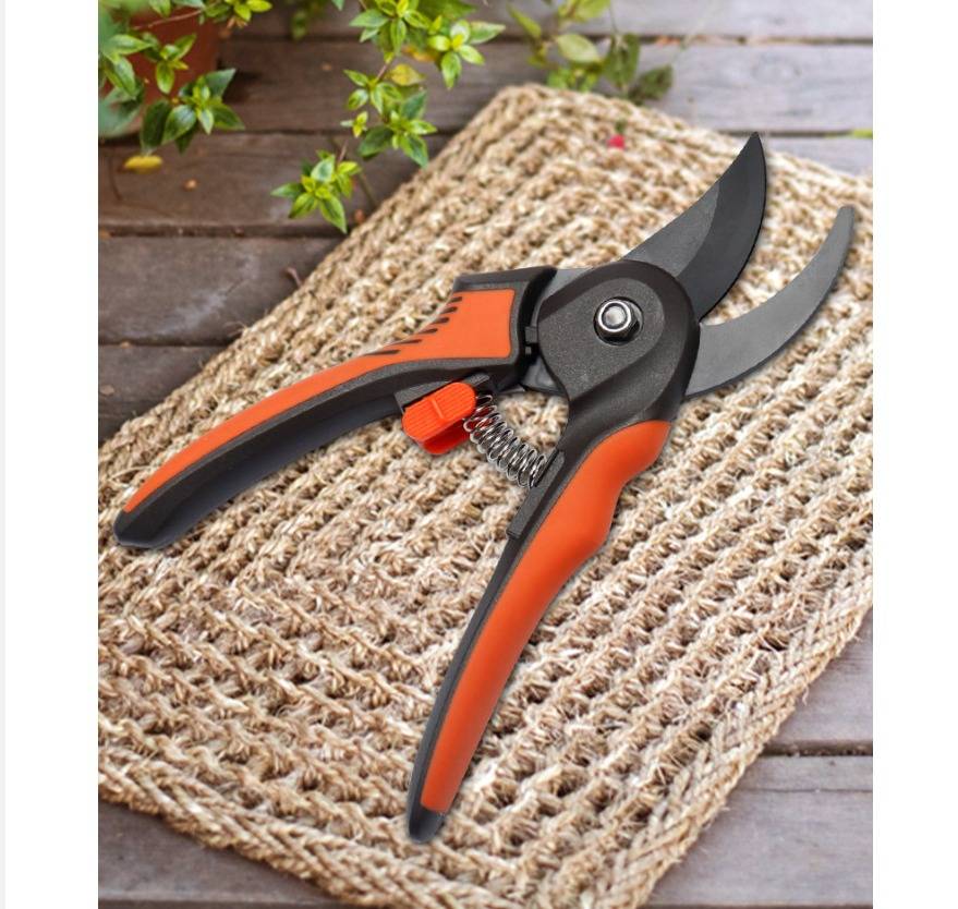 Offene Gartenschere mit orange-schwarzen Griffen, die auf einer Flechtmatte auf einer Holzoberfläche ruht, mit grünen Blättern in der Nähe.