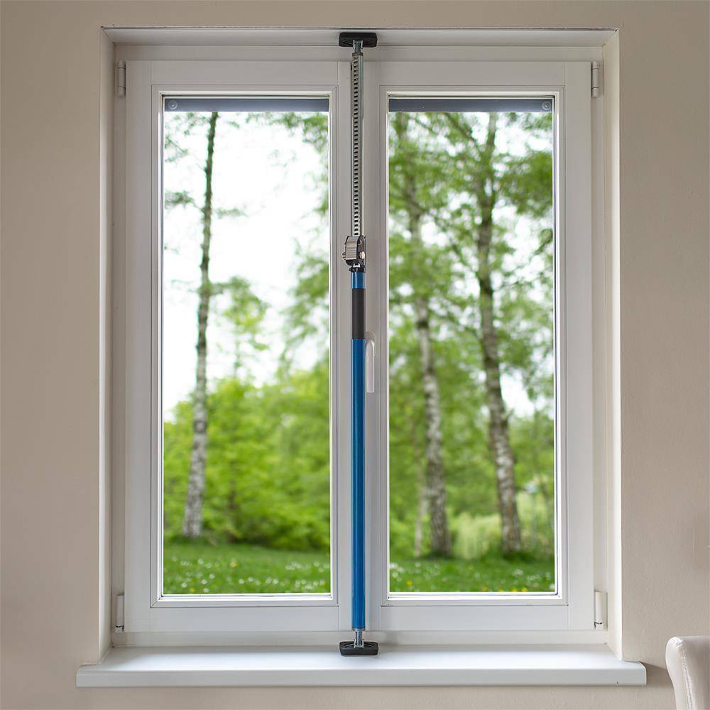 Ein blaues Reinigungswerkzeug hängt vertikal zwischen zwei geschlossenen weißen Fensterscheiben, mit grüner Waldaussicht draußen.