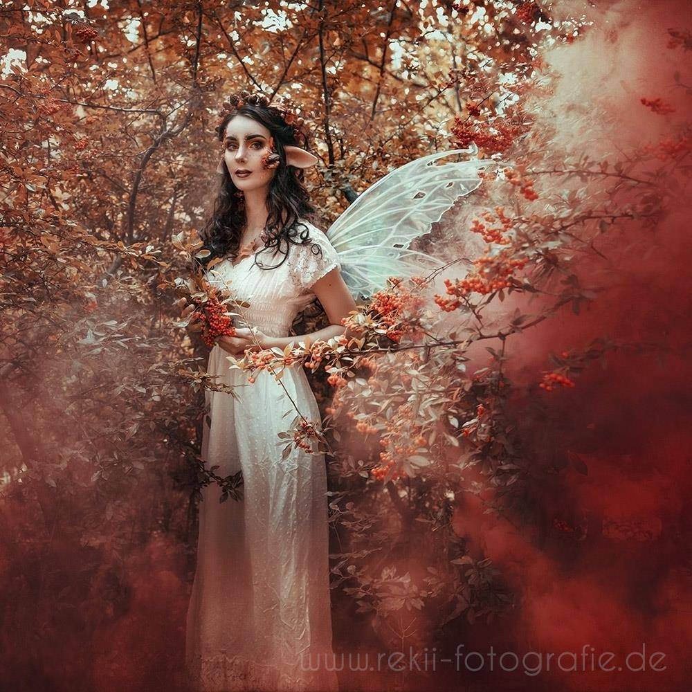 Eine elfenhafte Frau in einem weißen Kleid und durchsichtigen Flügeln steht inmitten von Herbstlaub mit rotem Nebel und hält Blumen in den Händen.
