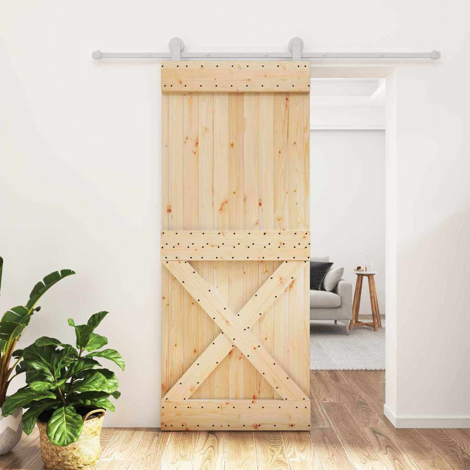 Türschieber im Scheunenstil aus Holz, der eine Türöffnung in einem hellen Wohnungsinneren teilweise abdeckt, mit einer Pflanze im Vordergrund und einem Blick auf ein Wohnzimmer dahinter.