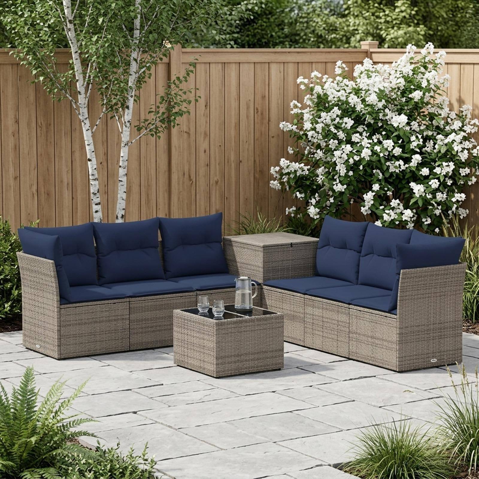 Außen-Terrassenbestuhlung mit zwei Korbbetten, Couchtisch und dunkelblau gefärbten Kissen auf einer Steinterrasse neben einem Holzzaun und blühenden Sträuchern.