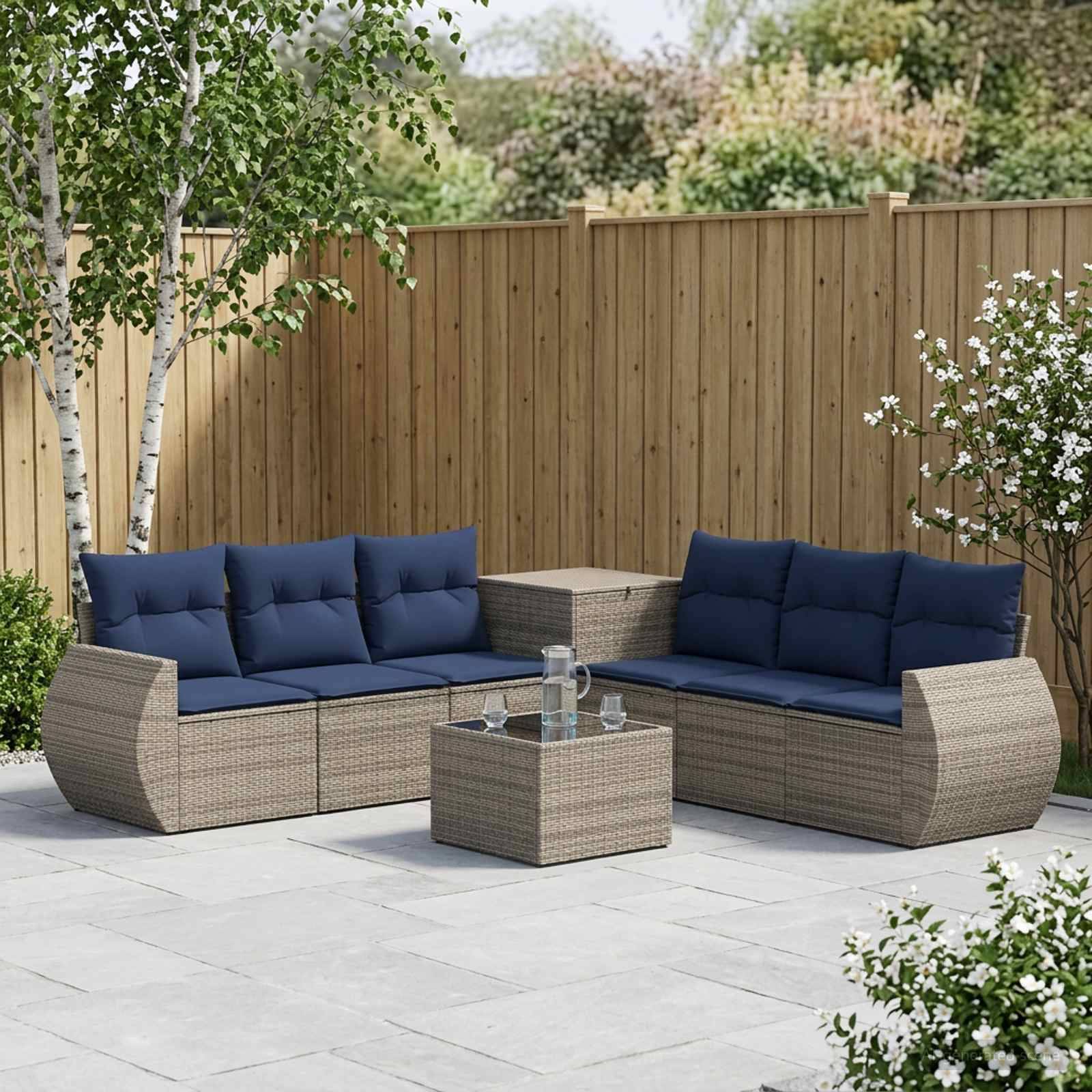 Zwei geflochtene Rattan-Außensofas mit marineblauen Kissen und ein passendes Tisch stehen auf einer Terrasse neben einem Holzzaun und blühenden Pflanzen.
