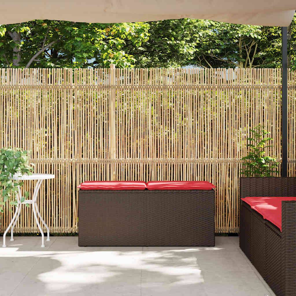 Terrassenfläche im Freien mit Bamboo-Sichtschutzwand, Korbmöbeln mit roten Kissen und einem kleinen weißen Beistelltisch im gesprenkelten Sonnenlicht.