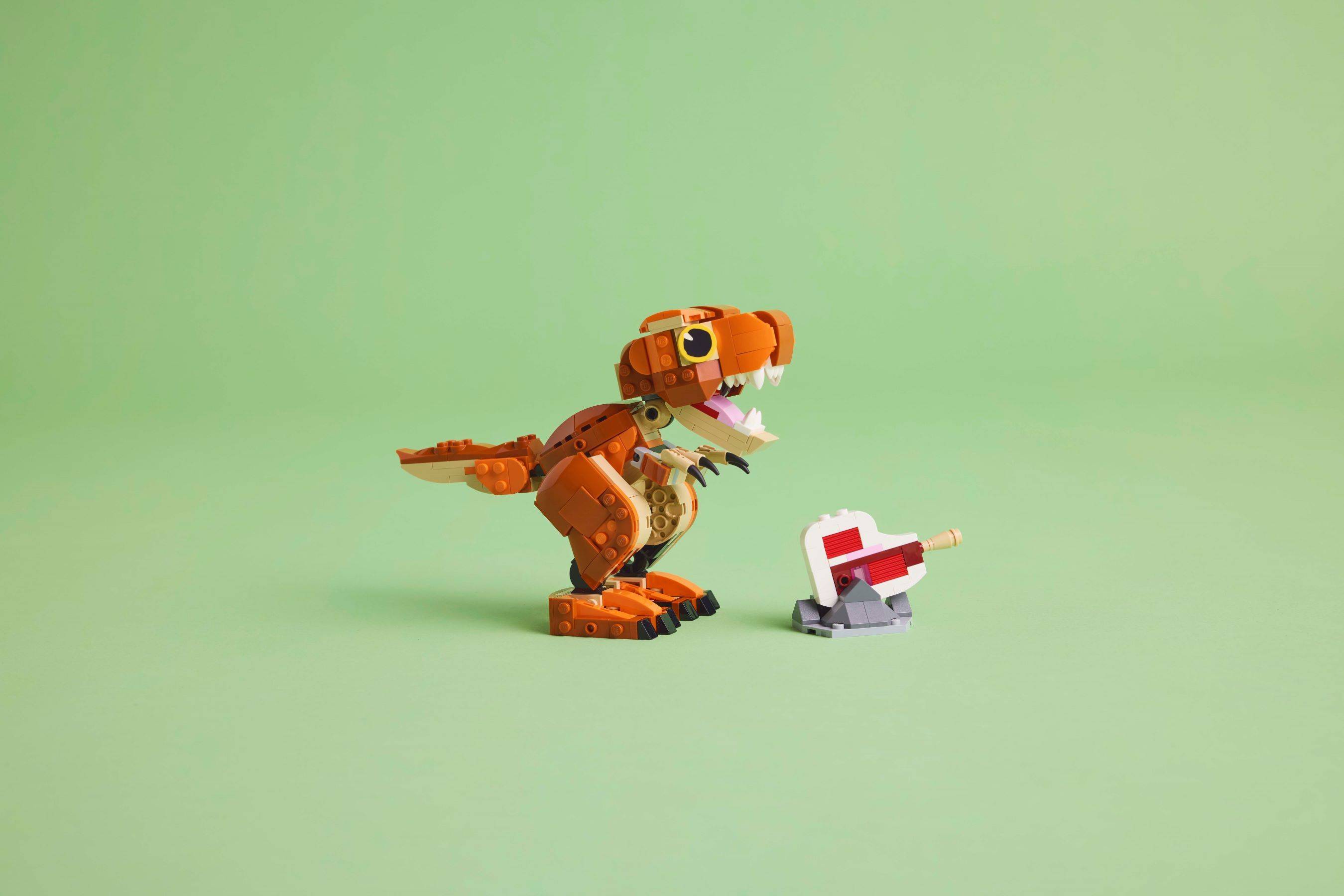 Orangefarbenes LEGO-Dinosaurier-Spielzeug, das auf eine kleine grau-rote Spielzeugpistole auf einfachem grünem Hintergrund gerichtet ist.