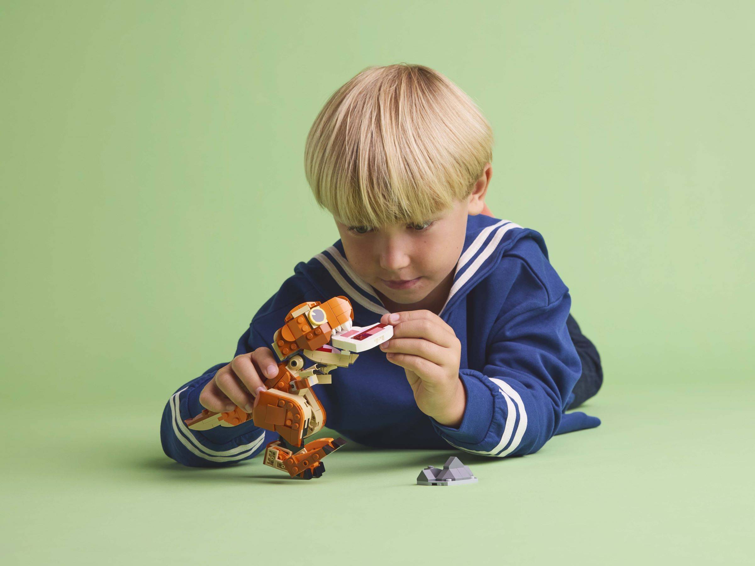 Ein kleiner Junge in einem blauen Kapuzenpullover liegt auf einem grünen Boden und montiert sorgfältig ein kleines orangefarbenes Roboterspielzeug.