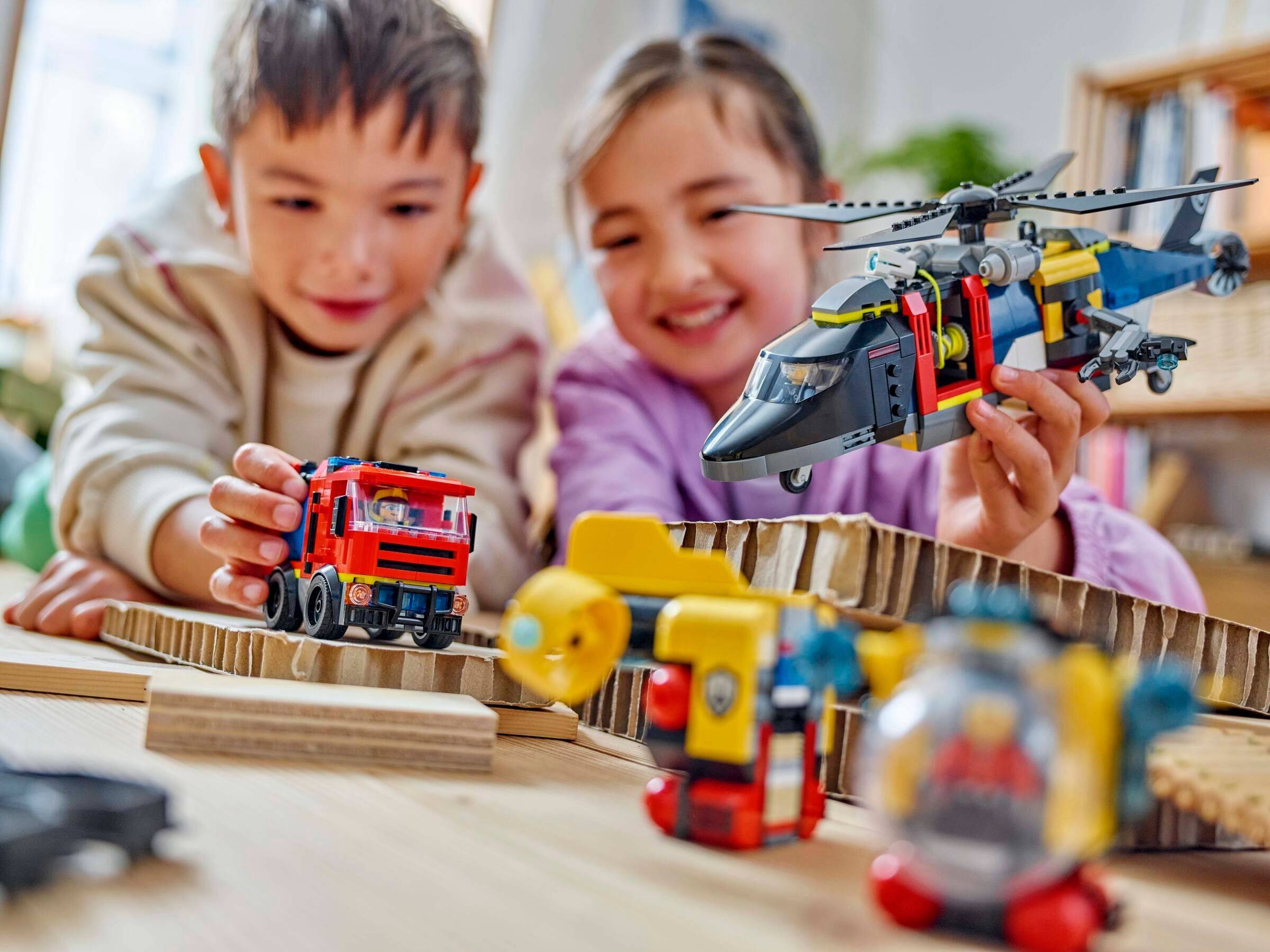 Zwei Kinder spielen mit Spielzeugfahrzeugen und einem Hubschrauber auf einer Pappstraße und lächeln drinnen an einem Tisch.