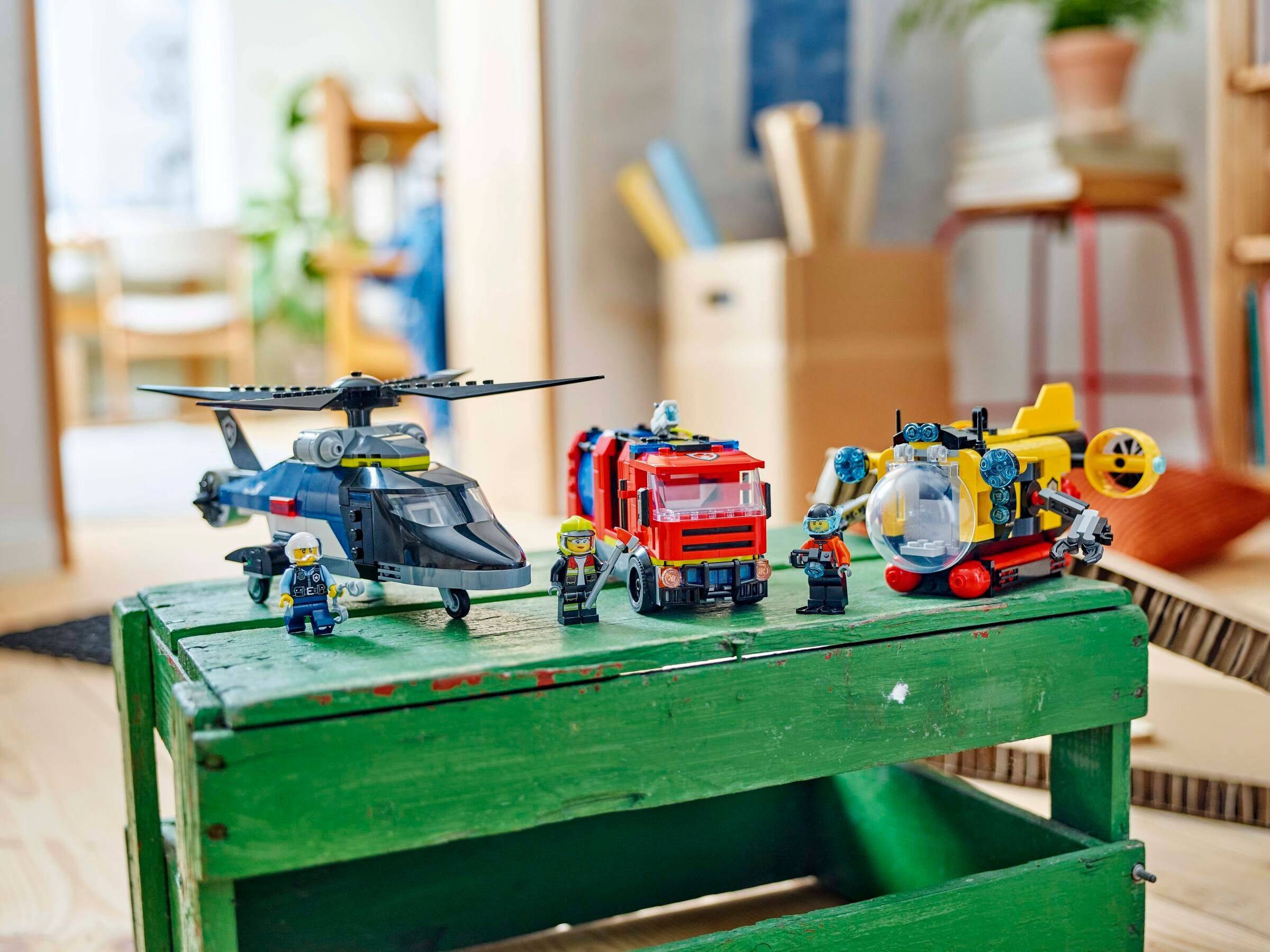 Lego-Notfallrettungsszenario mit einem Hubschrauber, einem Feuerwehrauto und einem Rettungsfahrzeug, jeweils mit Minifiguren, angeordnet auf einer grünen Holzkiste in Innenräumen.