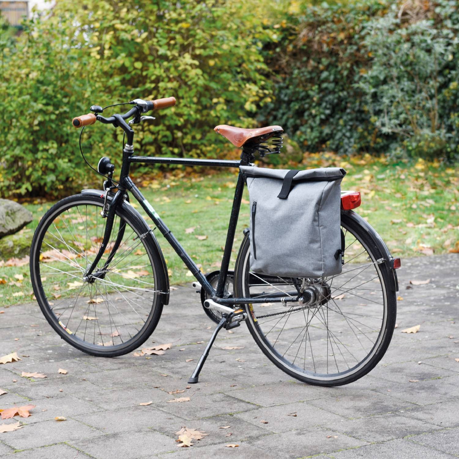 Schwarzes Pendlerfahrrad mit braunem Sattel und grauer Packtasche, geparkt auf einem gepflasterten Weg in einem parkähnlichen Gelände mit Baumbestand.