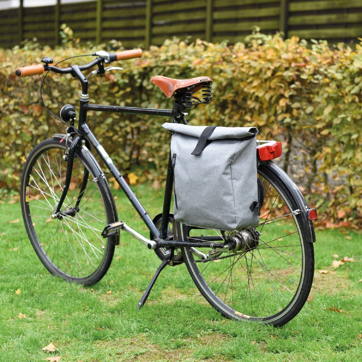 Schwarzes Fahrrad mit beigen Griffen und braunem Sattel, auf Gras geparkt mit einer grauen Hinterradtasche vor einer Hecke.