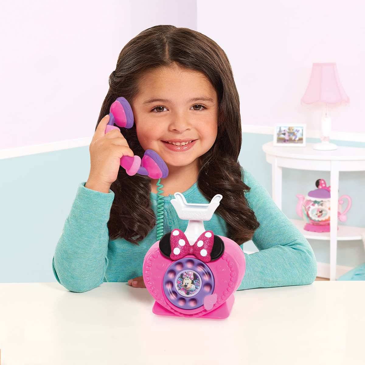 Ein lächelndes Mädchen hält ein rosa Spielzeug-Wählscheibentelefon mit einem violetten Hörer in einem Pastellzimmer.