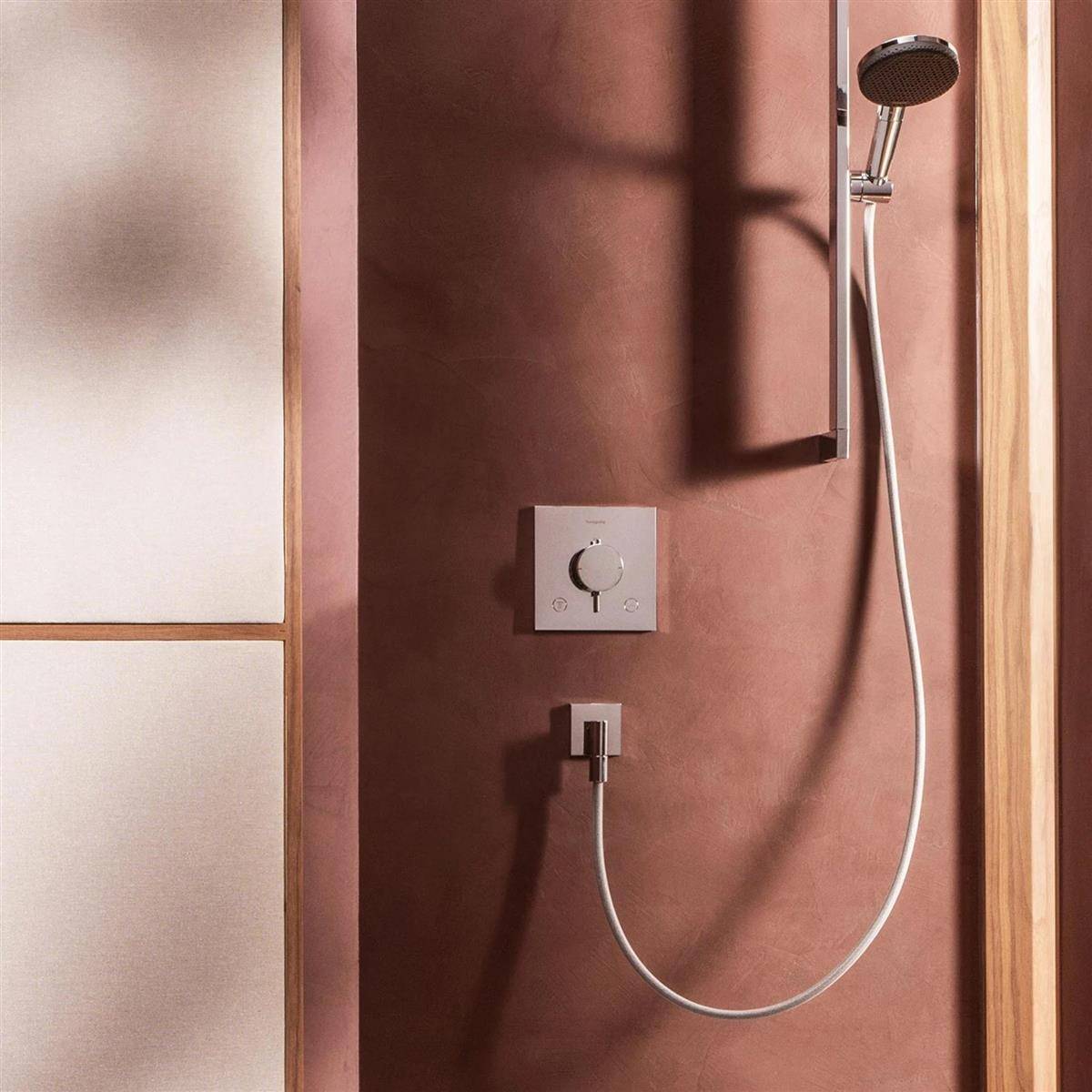 Minimale Duschfläche mit tragbarem Duschkopf, Kontrollregler und wandmontiertem Schlauch an einer braunen Kachelwand neben einer Milchglasplatte.