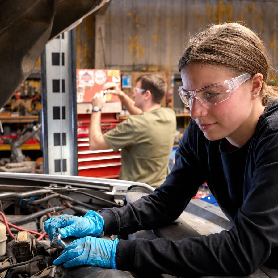 Eine Frau, die eine Schutzbrille und blaue Handschuhe trägt, arbeitet in einer Werkstatt an einem Automotor, während ein Mann im Hintergrund Werkzeuge organisiert.