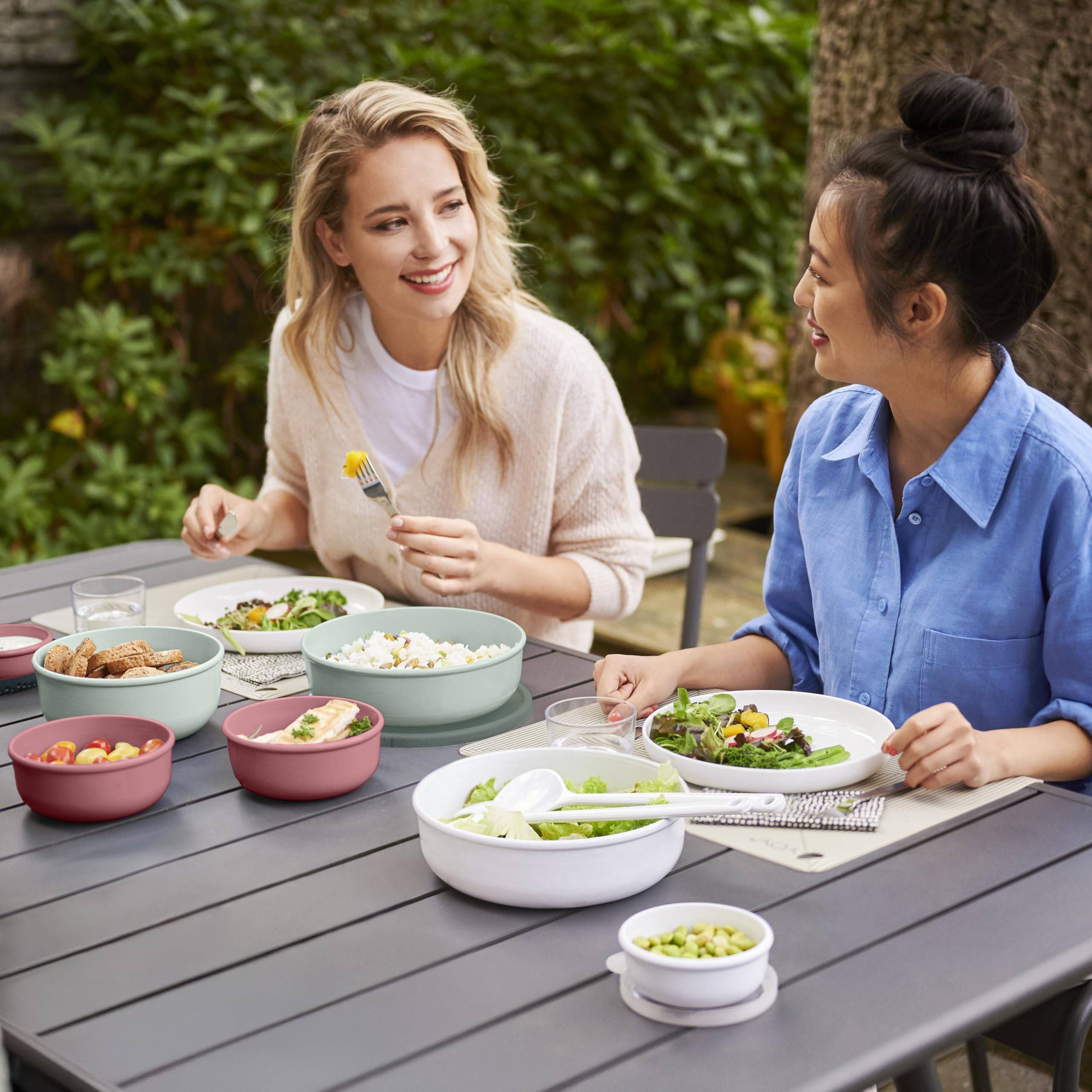 Zwei Frauen lächeln und unterhalten sich, während sie eine Mahlzeit im Freien an einem Tisch auf der Terrasse teilen, mit Schüsseln voller Salat und Beilagen.